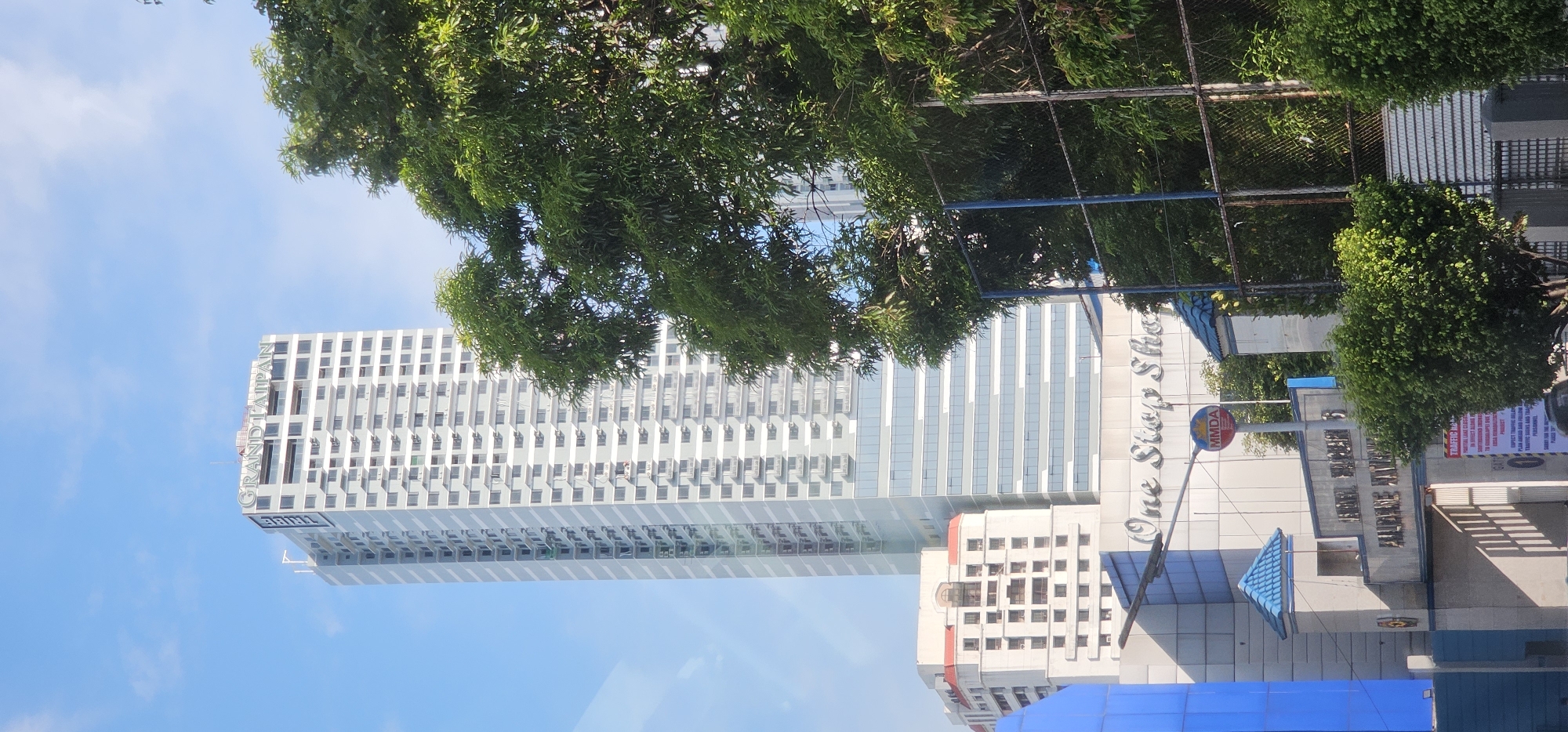 The image shows a bright, sunny day in Quezon City, Philippines, with a tall, modern skyscraper dominating the skyline. The skyscraper, identified by the text "GRAND TAIPAN" on its facade, appears to be a residential or commercial building with numerous windows and balconies. In the foreground, there is a lower building with a sign that reads "One Stop Shop," and to the right, a chain-link fence and lush green trees partially obscure the view. There are no people visible in the image. The sky is clear and blue, indicating good weather conditions. The overall scene depicts an urban environment in a commercial or mixed-use area.