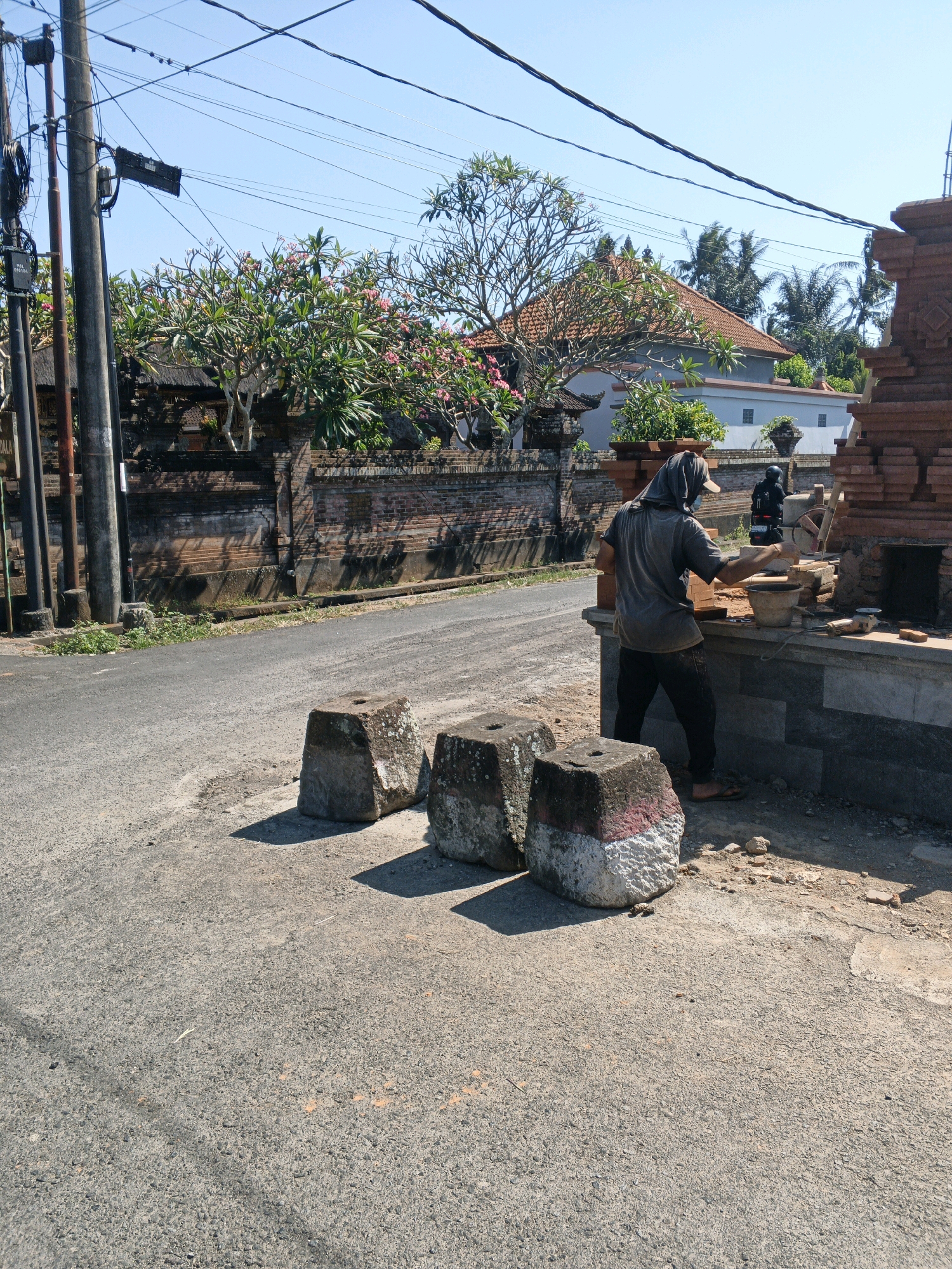 In Abiansemal, Indonesia, on a sunlit day, a construction worker is seen from the side, wearing a grey long-sleeved shirt and dark pants, with a head covering and mask. They are standing on the right side of the frame, actively engaged in building a brick structure. In front of them is a workbench holding various construction materials, including bricks, a bucket, a trowel, and a small scale. The worker is holding a brick in their right hand, suggesting they are in the process of laying it. A motorcycle with a rider in a black helmet is visible in the background, positioned behind the worker.
The immediate foreground features three weathered, square stone blocks with red and white paint markings on their sides, placed on the asphalt road. The road curves to the left, bordered by a traditional brick wall adorned with plants and flowers, a common sight in Balinese architecture. Beyond the wall, a house with a terracotta tiled roof and white walls is visible, along with palm trees and other tropical foliage under a clear blue sky. Utility poles with numerous wires stretch across the upper portion of the image, indicating the presence of electrical infrastructure. The scene depicts a typical street in Abiansemal, where construction work is taking place alongside residential and natural elements.