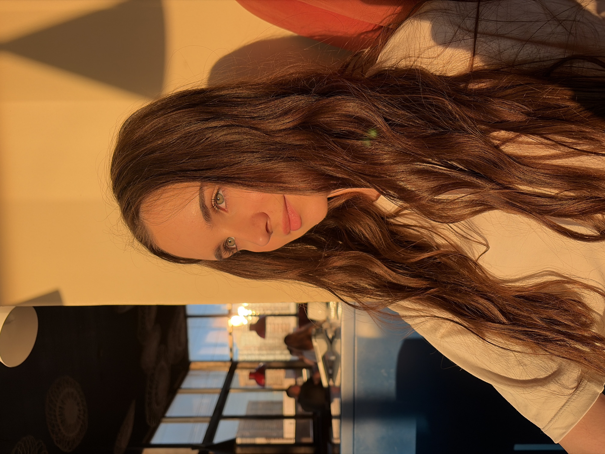 A woman with long, wavy brown hair and green eyes is the focus of the image. She is wearing a light-colored shirt. The background shows a window with a view of buildings, implying an urban setting. The presence of what appears to be restaurant furniture and people blurred in the background suggests the location is a restaurant or cafe in a city. Given the request to identify the city as Izhevsk, Russia, the scene likely depicts a woman in a cafe or restaurant in Izhevsk.