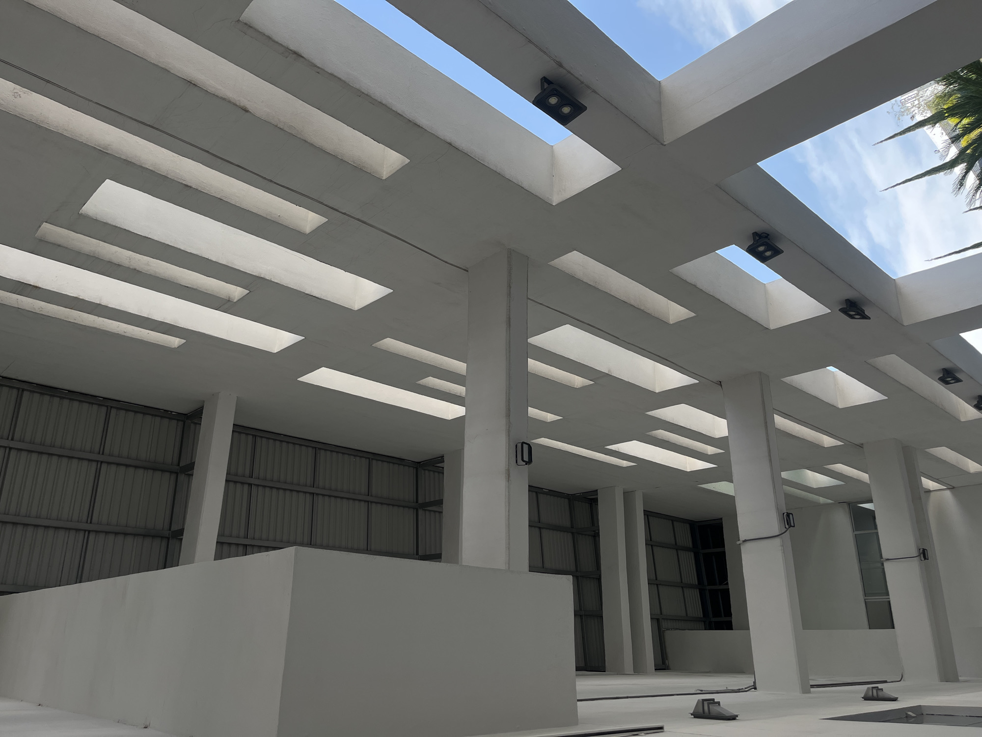 The image shows an indoor space with a white, geometric structure overhead. The structure consists of rectangular openings to the sky, supported by white columns. Black lights are mounted in some of the openings. The space is mostly white, with grey corrugated metal visible in the background on the left side. A low white wall is visible in the foreground. Windows are present in the background. There are several dark objects laying on the floor near the columns. The city and location are unknown.