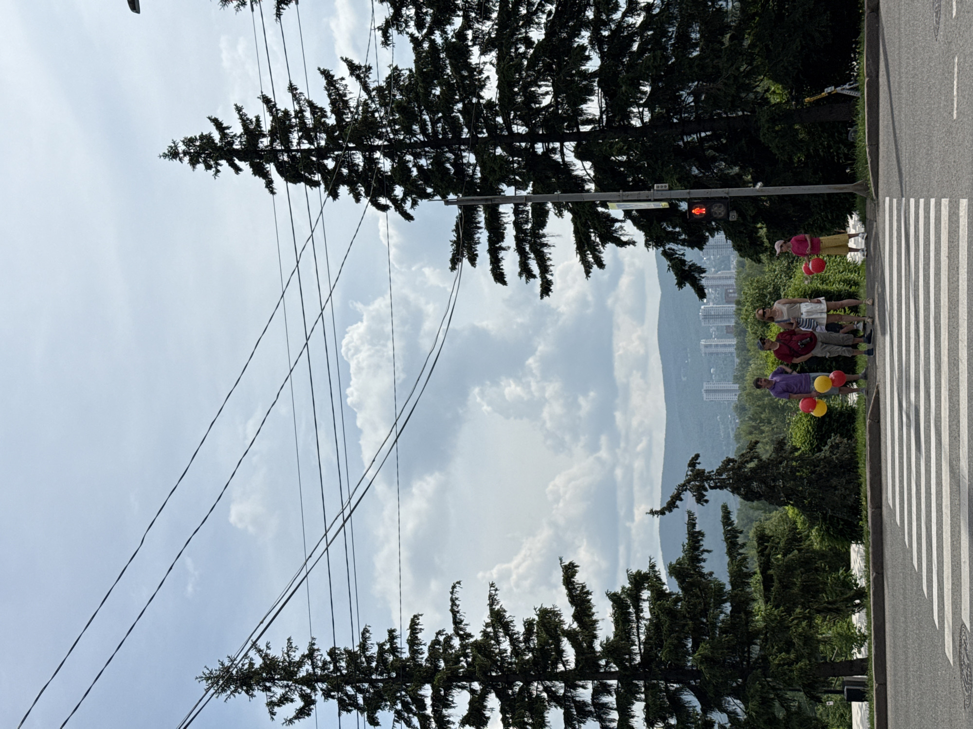 In Krasnoyarsk, Russia, a group of five people stand near a crosswalk, some holding red and yellow balloons. A traffic light shows red for pedestrians. Towering evergreen trees frame the scene, with power lines stretching across the sky. In the background, distant hills overlook a cityscape with modern buildings. The weather appears to be partly cloudy.