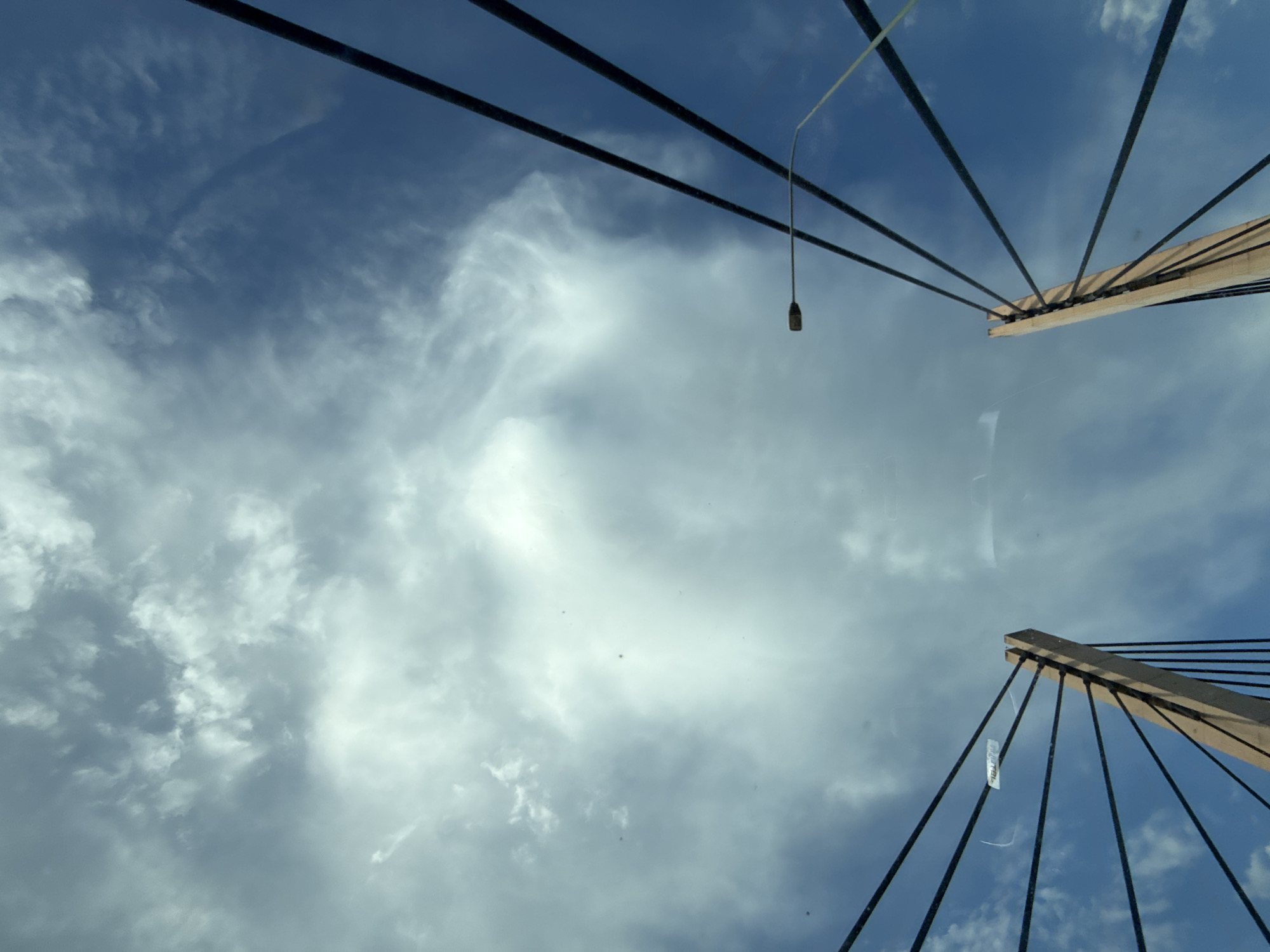 The image is a low-angle shot of a suspension bridge against a cloudy blue sky. The bridge's beige-colored concrete towers are visible on the left and right sides, with dark cables extending upwards and outwards. A streetlamp is positioned on the right side of the frame. The clouds are dense and white in the upper portion of the image, with areas of clear blue sky below. A small white tag with some text is attached to one of the cables on the left. The scene appears to be captured from beneath the bridge, looking upwards. The specific location in Wrocław, Poland is not determinable from the visual information alone.