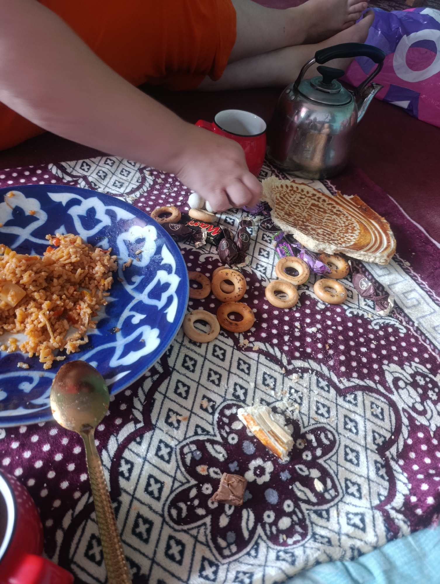 The image depicts a domestic scene on a patterned fabric surface, likely in Marhamat, Uzbekistan. A person's arm, partially visible and wearing an orange garment, is reaching towards a blue and white patterned plate containing a serving of pilaf, or plov. Next to the plate are several small, ring-shaped baked goods, possibly kalachi or similar bread rings, and wrapped candies, including one appearing to be a Mars bar. A silver spoon with a textured handle rests on the fabric near the plate. A red mug sits on the fabric to the left of a metal kettle with a black handle. In the background, legs of a person are visible, and a purple and white patterned shopping bag is partially seen. The overall environment suggests a casual meal or snack time in a home setting.