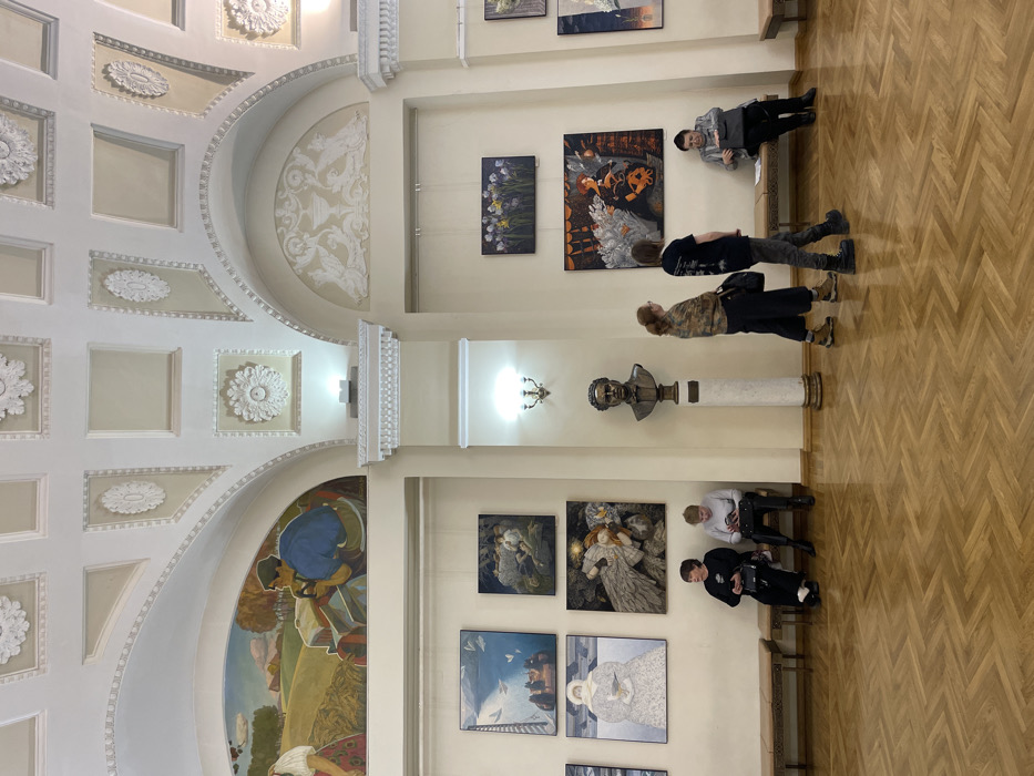 The image captures an expansive, well-lit interior hall within the Yekaterinburg Museum of Fine Arts in Yekaterinburg, Russia. The hall features high, light-colored walls with prominent architectural arches and a richly detailed ceiling adorned with decorative white plaster moldings and rosettes. A large, vibrant mural depicting a rural landscape with a person on a tractor is visible within an arch on the upper left, while a white ornamental plaster relief, featuring a central urn and two winged creatures, occupies an arch on the upper right.
The walls are furnished with numerous framed paintings of various sizes, hung by visible wires. On the left wall, a cluster of five paintings includes scenes with blue skies, figures in darker settings, and a person in a white coat holding a bird. On the right wall, a painting of purple irises and a larger artwork featuring a figure with white, wing-like elements are prominent. Centrally located, a bronze bust of a man with short, wavy hair is mounted on a tall, white fluted column with a square base. The floor is covered with a polished wooden parquet laid in a herringbone pattern.
Six individuals are present in the scene. On a wooden bench along the left wall, two women are seated: one in a black top holding a handbag, and another in a light top focused on a smartphone. A third person is partially visible on a bench further left, also looking at a device. In the mid-ground, two individuals stand, facing right; one with reddish-brown hair wears a patterned jacket, while the other, with dark hair and a black t-shirt, carries a black bag. On a bench against the right wall, a man in a dark jacket is seated, looking down at an object in his hands. The hall is illuminated by several wall sconces with multiple bulbs and ambient lighting.