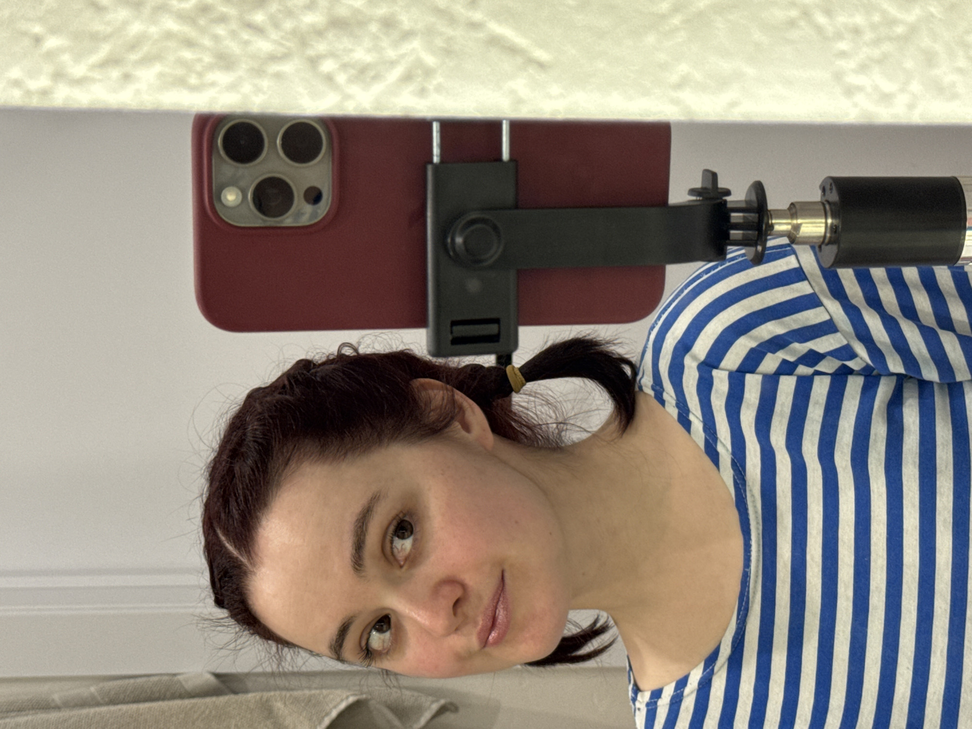 The image displays a young woman looking at the camera, with a smartphone mounted above her head. The photo is rotated clockwise, showing the woman's head tilted towards the bottom of the frame and the phone on the left side.
The primary subject is a young woman with dark hair, which appears to be styled into small pigtails or braids held by a light-colored elastic band. She has light skin, brown eyes, and is looking directly towards the viewer with a gentle, slightly smiling expression. She is wearing a sleeveless top with prominent vertical blue and white stripes.
Above and slightly to the left of her head, a dark red smartphone, consistent with an iPhone model featuring a triple camera array, is securely held by a black phone mount. This mount is attached to a metallic rod or arm, suggesting it's part of a larger stand or tripod, positioning the phone to face the woman.
The setting appears to be indoors. Behind the woman and the phone is a plain, light-colored wall. Above the phone, a textured, off-white surface, likely a ceiling or upper wall, is visible.
The activity depicted suggests the woman is either taking a selfie, recording a video, or participating in a video call, utilizing the mounted phone for a clear shot. There is no visible text in the image. While the provided context specifies Izhevsk, Russia, no specific visual cues within the image confirm this location. The overall mood is calm.