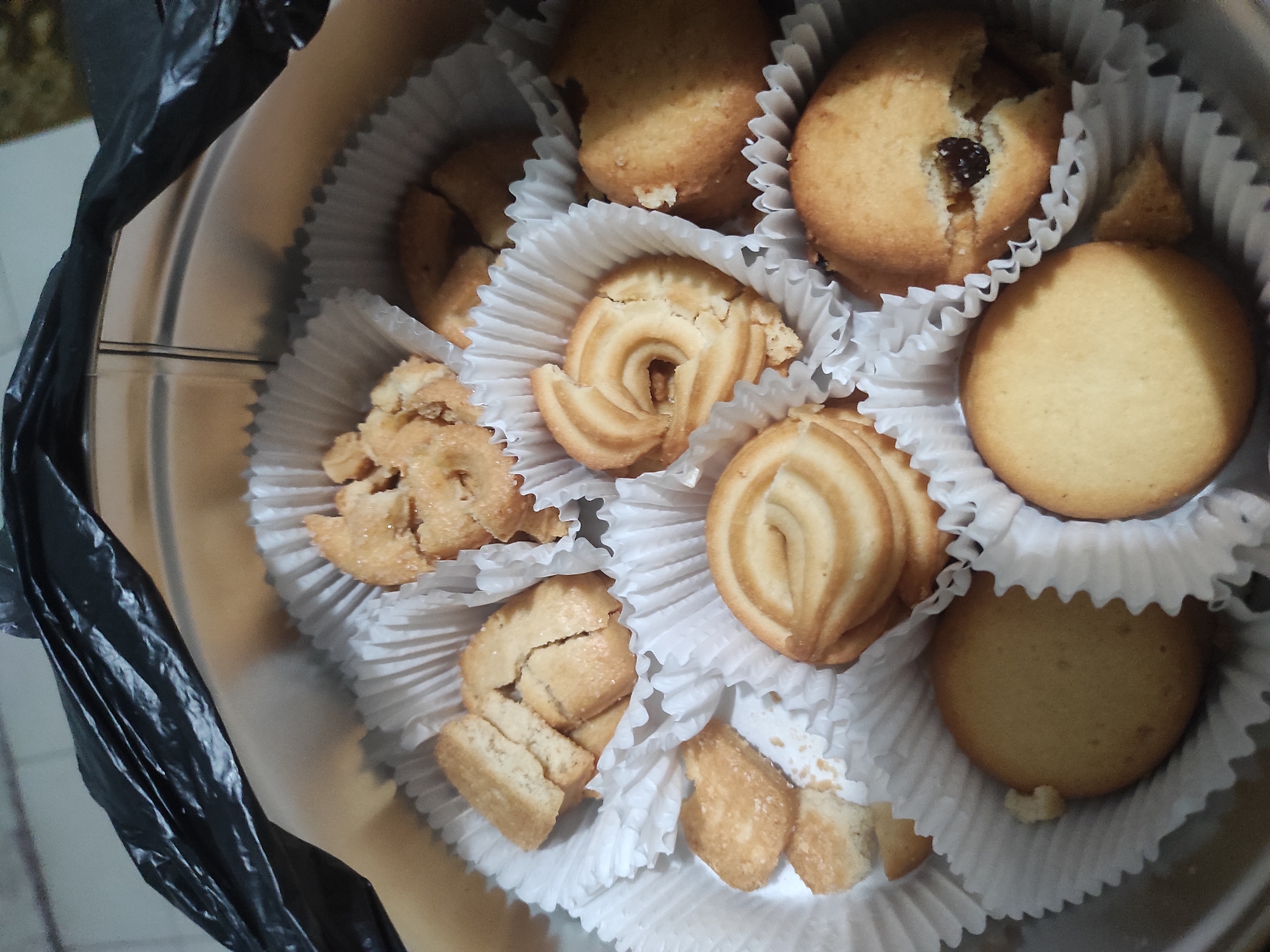 A close-up, overhead shot shows an assortment of cookies in a round metal tin, lined with white paper cupcake liners. The cookies vary in shape and texture. Some are golden brown and smooth, while others are a lighter tan and appear to be crumbled or broken. One cookie has a swirl pattern, and another has a dark raisin embedded in it. The tin is partially covered by a crinkled black plastic liner. The background is blurred but appears to be a countertop with a white tiled surface. The image does not contain any elements that would allow for determination of its location in Sumedang, Indonesia.