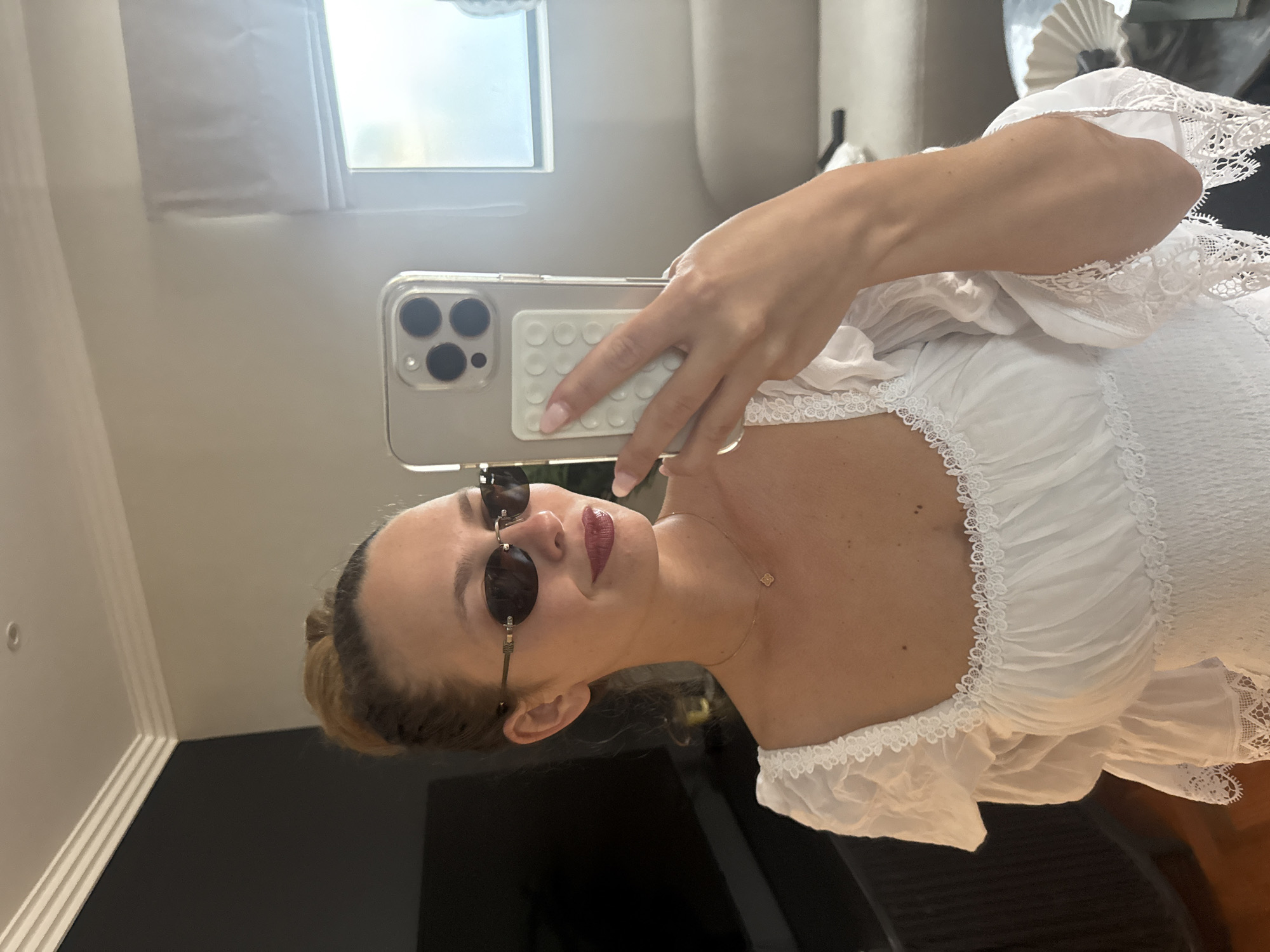 A woman is taking a selfie indoors in a room with neutral-colored walls. She is wearing sunglasses and a white, frilly top. She is holding a smartphone with a gray case in her right hand. A window is in the background to the left, and dark furniture is visible below. The location cannot be determined as Cape Town, South Africa from the given visual information.