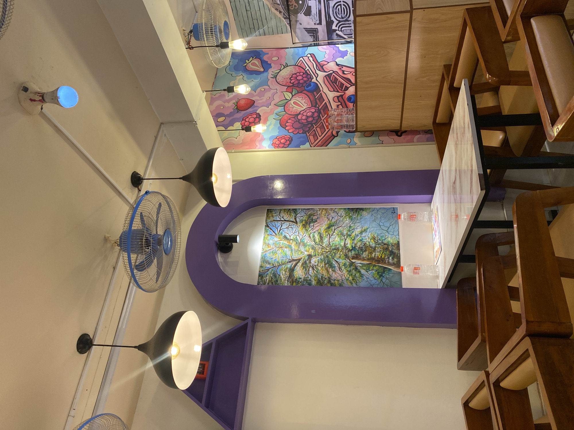 The image shows the interior of a business, possibly a cafe or restaurant in Dhaka, Bangladesh. The ceiling features a decorative mural with a whimsical strawberry waffle design and a painting of trees framed by a large purple arch. Blue electric fans and black pendant lights are also mounted on the ceiling. A table is set with water bottles. Wooden chairs are stacked nearby.