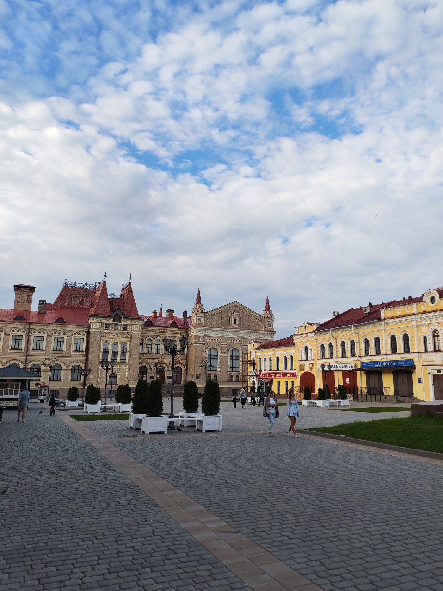 The image depicts a street scene in Bol'shie Mkhi, Russia, featuring historic buildings and a paved walkway. The architecture includes a prominent building with a red-tiled roof and ornate details, juxtaposed with a bright yellow building housing shops. Several people are visible walking and one person is on a bicycle. The ground is covered in grey cobblestones, with white planters containing green trees. The sky is partly cloudy, with a mixture of blue sky and white clouds. The overall impression is of a public square in a historic part of a Russian city.