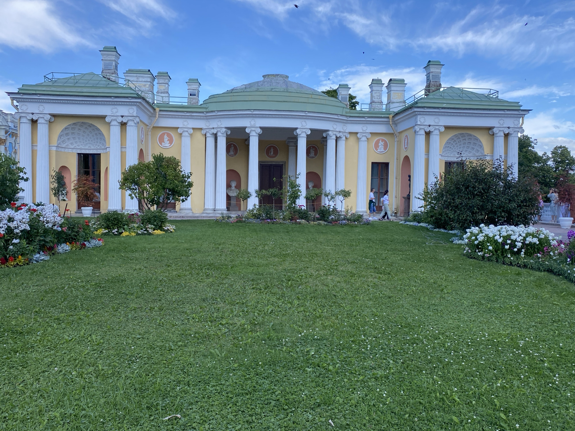 This image depicts the Chinese Palace in the Catherine Park, city of Pushkin, Russia, under a partly cloudy sky. The palace, painted a pale yellow, features a prominent semicircular portico supported by numerous white classical columns. Green roofs top the structure, with decorative chimneys. Circular medallions with relief figures adorn the facade. A well-maintained green lawn stretches out in front of the palace, bordered by flowerbeds with colorful blooms, including white, red, and yellow flowers. A few trees and bushes are also visible, contributing to the park-like setting. Several people are present, walking and standing near the palace. One person is walking towards the right side of the frame, while two others are seen near the entrance. A few birds are flying in the sky. The overall scene conveys a sense of serene parkland with historical architecture.