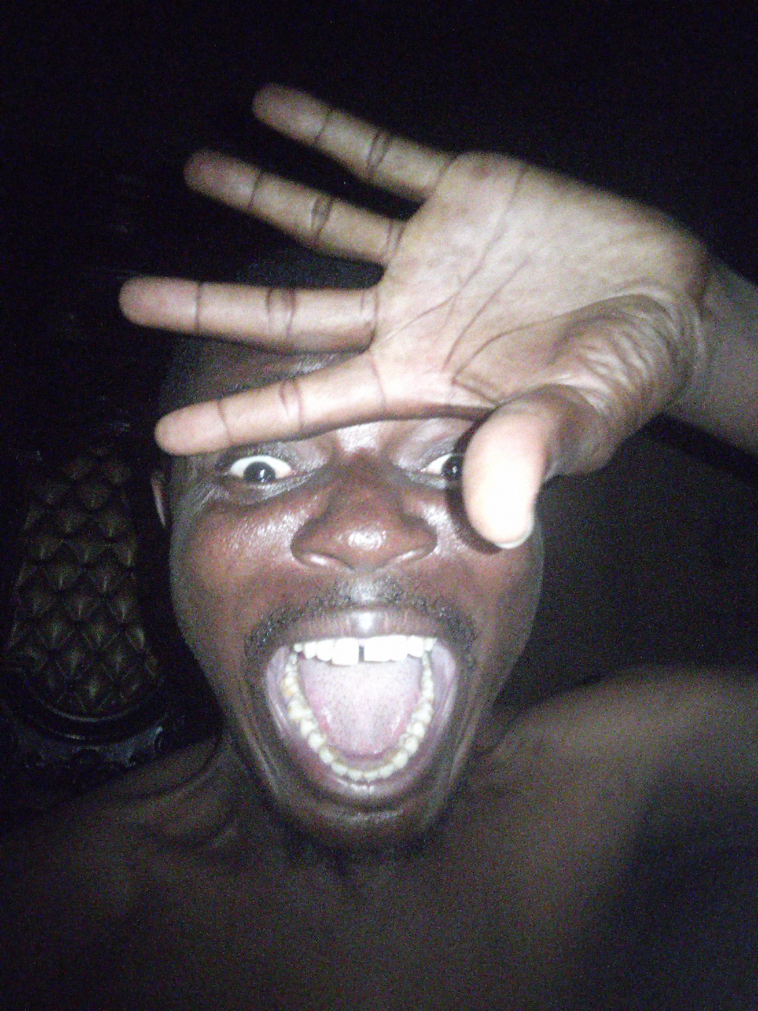 A close-up, low-light, grainy photo shows a Black man with his mouth wide open and his eyes open, looking at the camera. His right hand is raised to his face, with his index finger in front of his right eye and his other fingers spread. The man's teeth are visible. The background is dark and blurry, with a patterned texture visible on the left side of the frame.