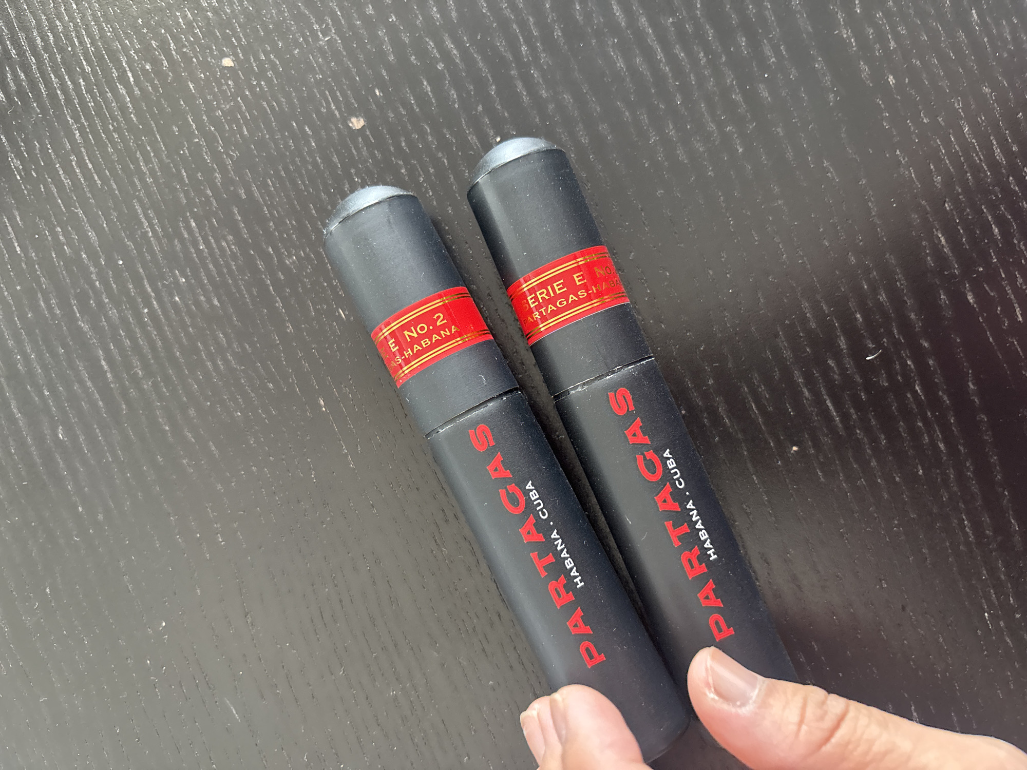Two black cigar tubes with red labeling are laid on a dark, textured surface. The top cigar tube has a red band with gold lettering that reads "SERIE E NO. 2 HABANA. CUBA". The bottom cigar tube also has a red band with gold lettering that reads "SERIE E N PARTAGAS HABANA, CUBA". The name "PARTAGAS" is printed in red lettering on both cigar tubes. A hand with visible fingernails is partially visible on the left side of the frame, with fingers touching the bottom cigar tube. The background surface appears to be a dark wooden table with a visible wood grain pattern and some dust or debris. The image was taken in Dubai, United Arab Emirates.
