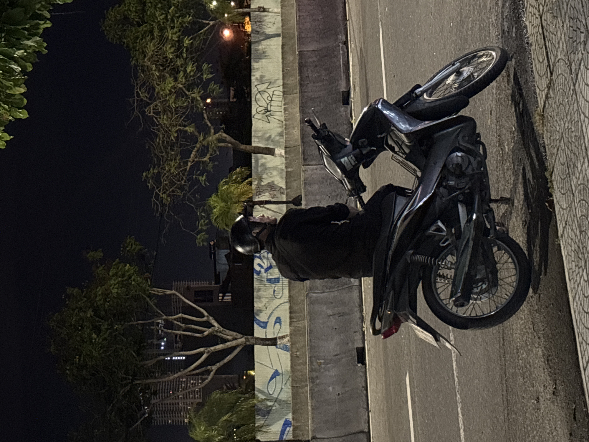 A person wearing a black shirt and black helmet sits on a black motorcycle. The motorcycle is parked on the side of a road. In the background, there is a concrete barrier with graffiti on it. Behind the barrier, there are trees and buildings. The scene appears to be at night. This scene is likely in Đà Nẵng, Vietnam, given the presence of motorcycles and the style of buildings and graffiti.