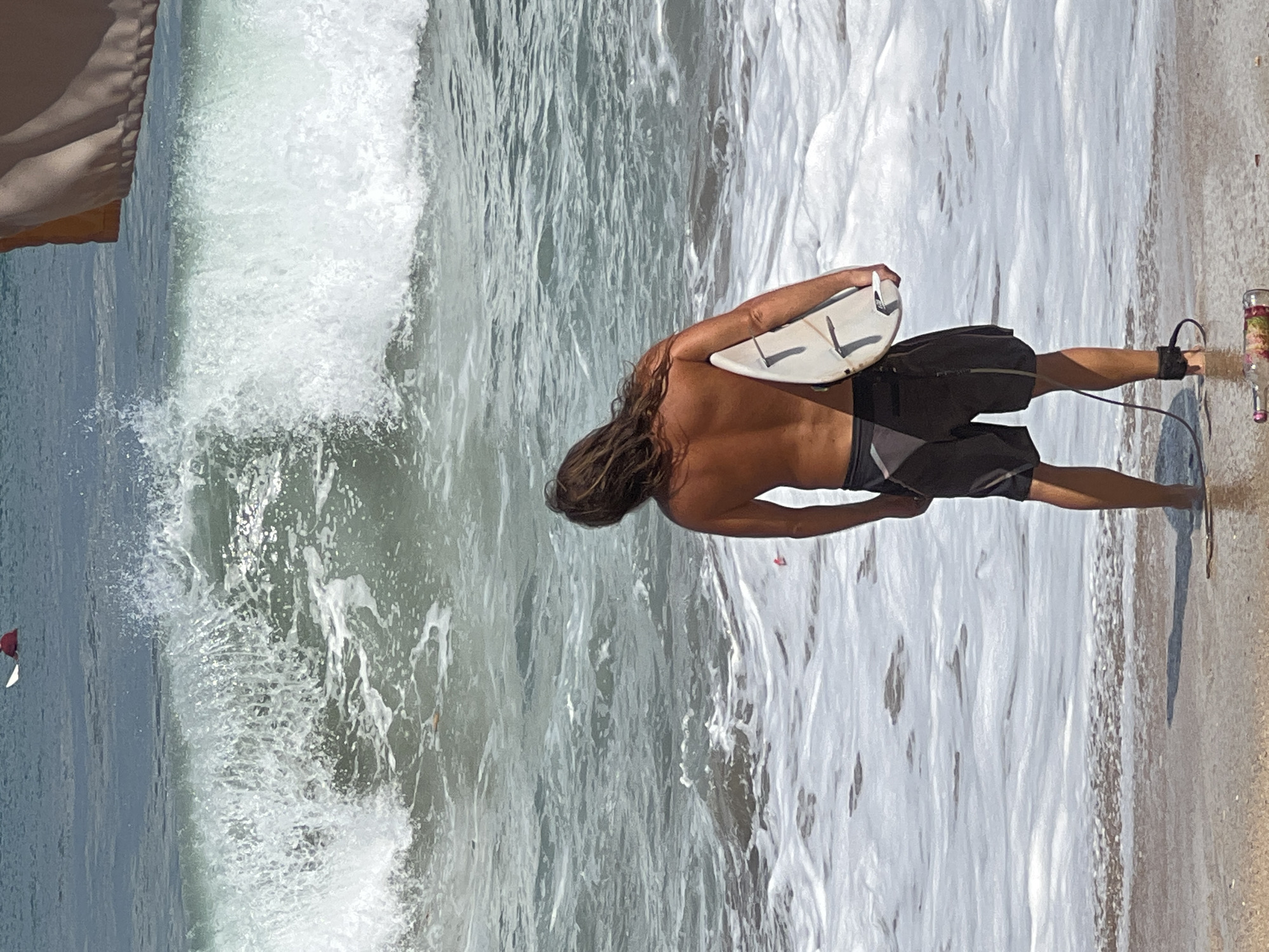 A shirtless surfer with long brown hair stands in shallow water at Kuta Beach, Indonesia, holding a white surfboard with black markings under his right arm. He wears black board shorts and an ankle leash. The surfer is facing away from the camera, looking towards a large wave breaking in the ocean. In the background, another surfer in a red top is visible further out in the water. The sand is visible in the foreground, and a glass bottle with a pink and floral label lies on the wet sand near the water's edge. The environment is a sunny beach with foamy waves and blue-grey water.