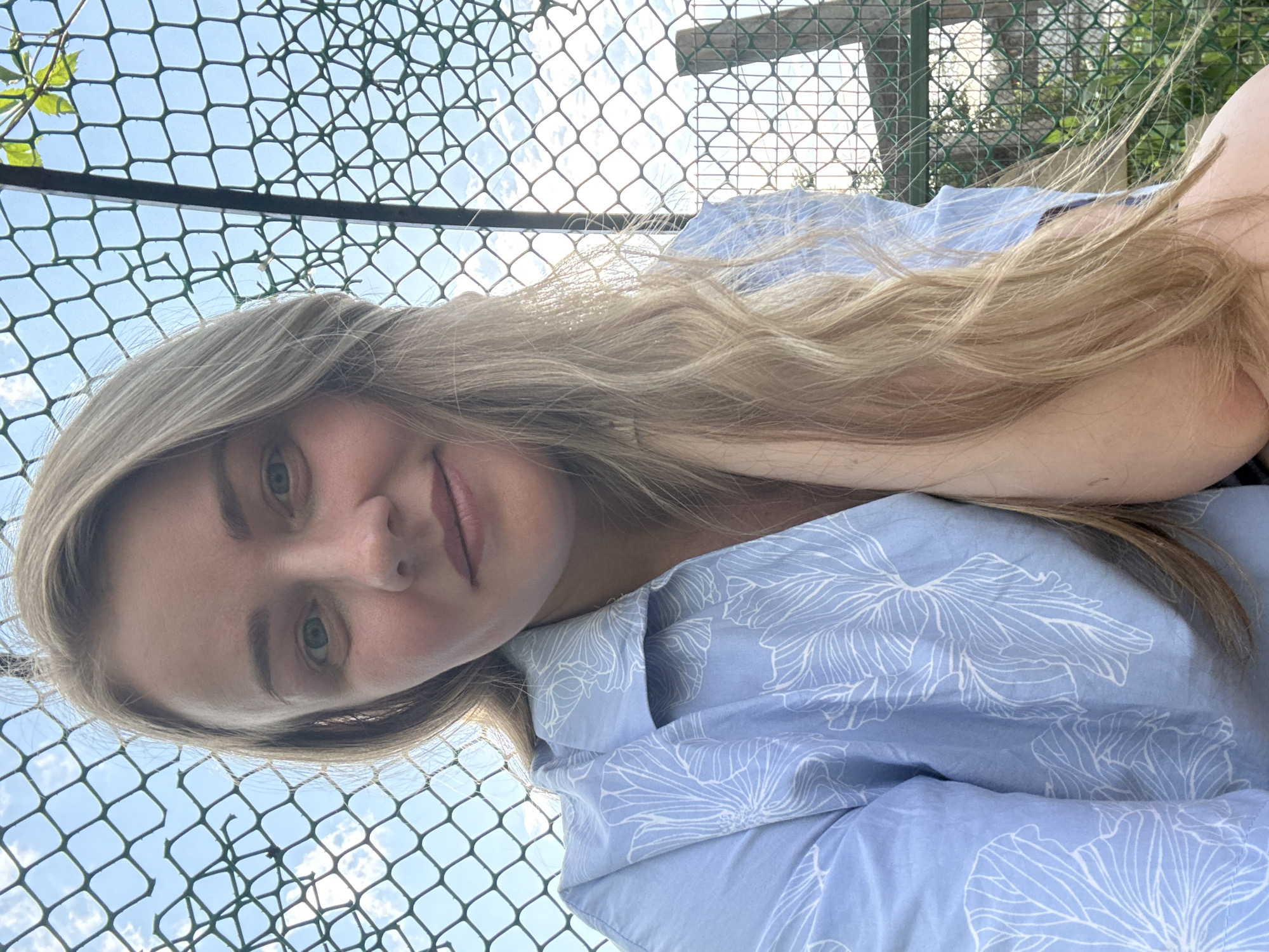 A woman with blonde hair and a blue floral-patterned shirt is looking at the camera. She is positioned against a backdrop of a mesh fence, with a glimpse of sky visible through the mesh. The location is unspecified.