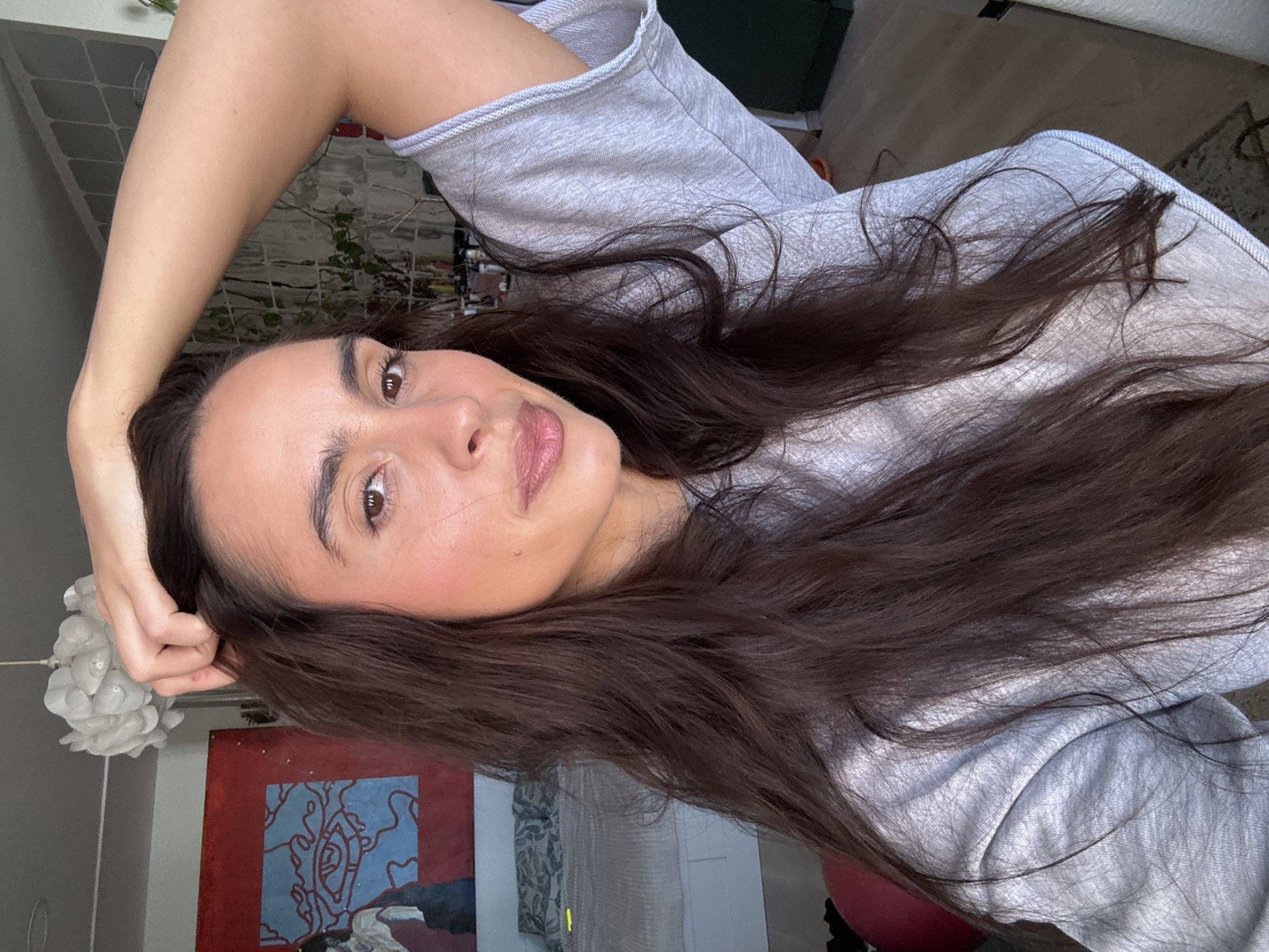 The image is a selfie of a woman with long, dark brown hair. She is wearing a light gray sweatshirt. Her left arm is raised and her hand is resting on her head. She has brown eyes and is looking at the camera. The background includes a painting with blue, red, and white colors, a white paper lantern, a gray wall, and a glimpse of greenery through a window. There is no information provided that suggests the image was taken in Krasnoyarsk, Russia.