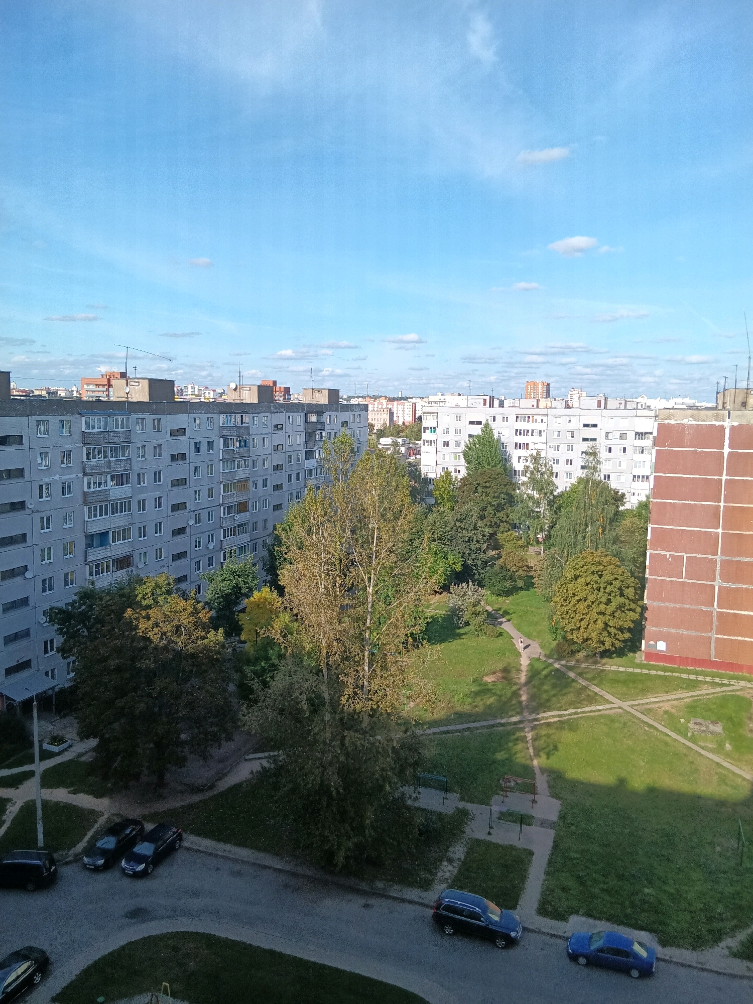 This image depicts a residential area in Mahilyow, Belarus, on a clear day with scattered clouds. The scene is dominated by a large, multi-story apartment building with a grey facade, multiple windows, and balconies. In the foreground, a paved road runs parallel to the apartment building, with several cars parked along its edge. These include a dark-colored SUV, two dark-colored sedans, and a blue sedan. Adjacent to the road and building is a green lawn interspersed with trees. A path cuts through the lawn, and a lone pedestrian is visible walking on it. Another tall building with a red brick facade and white trim is situated to the right of the frame, partially obscuring the view of other structures in the background. The background reveals a cityscape with more apartment buildings of varying heights and architectural styles. The overall impression is a typical urban residential environment.