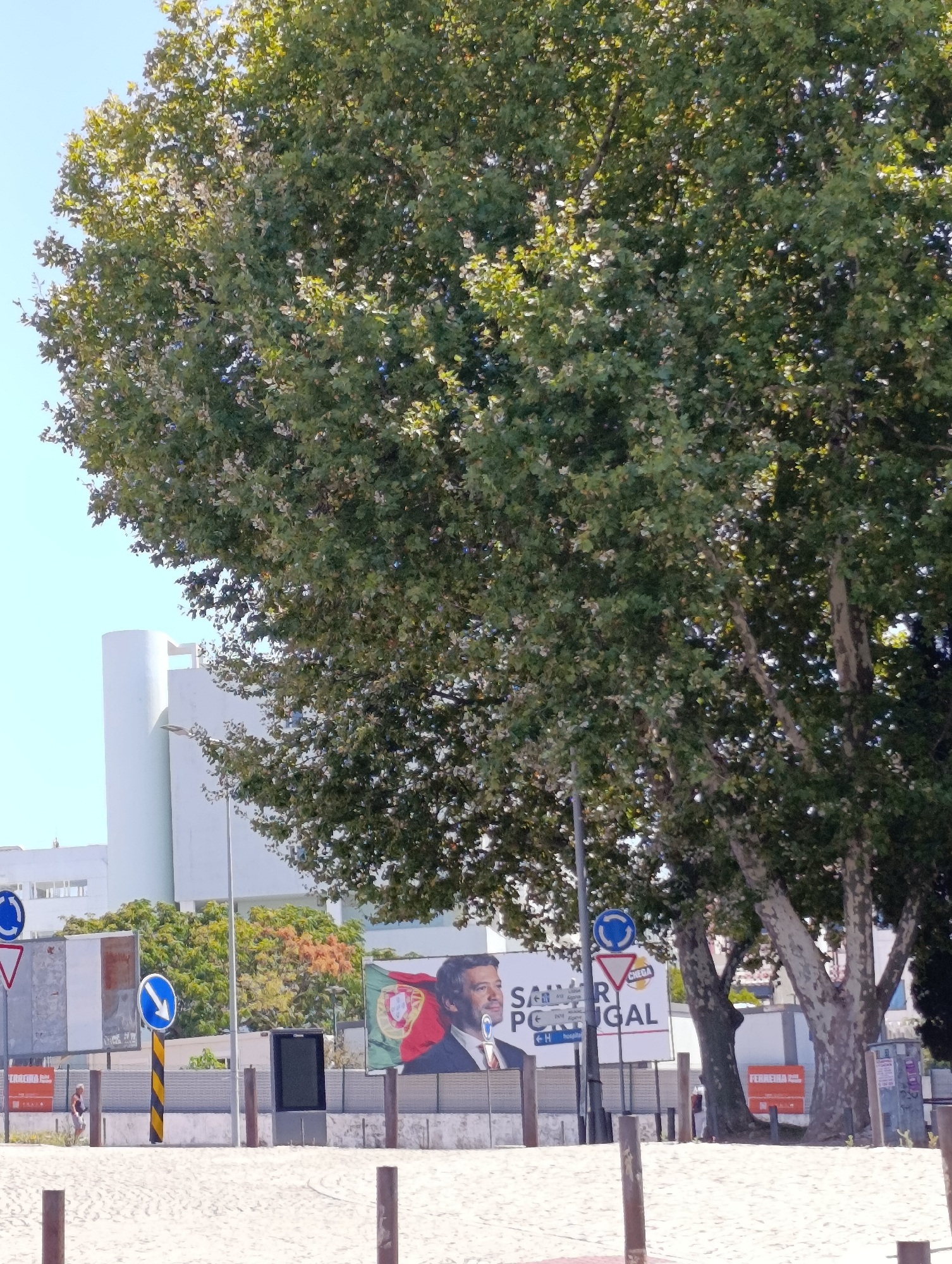A sunlit exterior shot shows a large tree in the foreground, its leaves dappled with sunlight. Behind the tree, a modern white building with cylindrical elements is visible. Several billboards are positioned behind a low wall. One prominent billboard features a portrait of a man, accompanied by text that includes the words "SALVAR PORTUGAL" (Save Portugal) and the Portuguese flag. Road signs, including a circular "Roundabout" sign and a directional arrow sign, are also present. A sandy ground is visible in the foreground, suggesting a beach or promenade area. The image is likely taken in Setúbal, Portugal, given the signage.