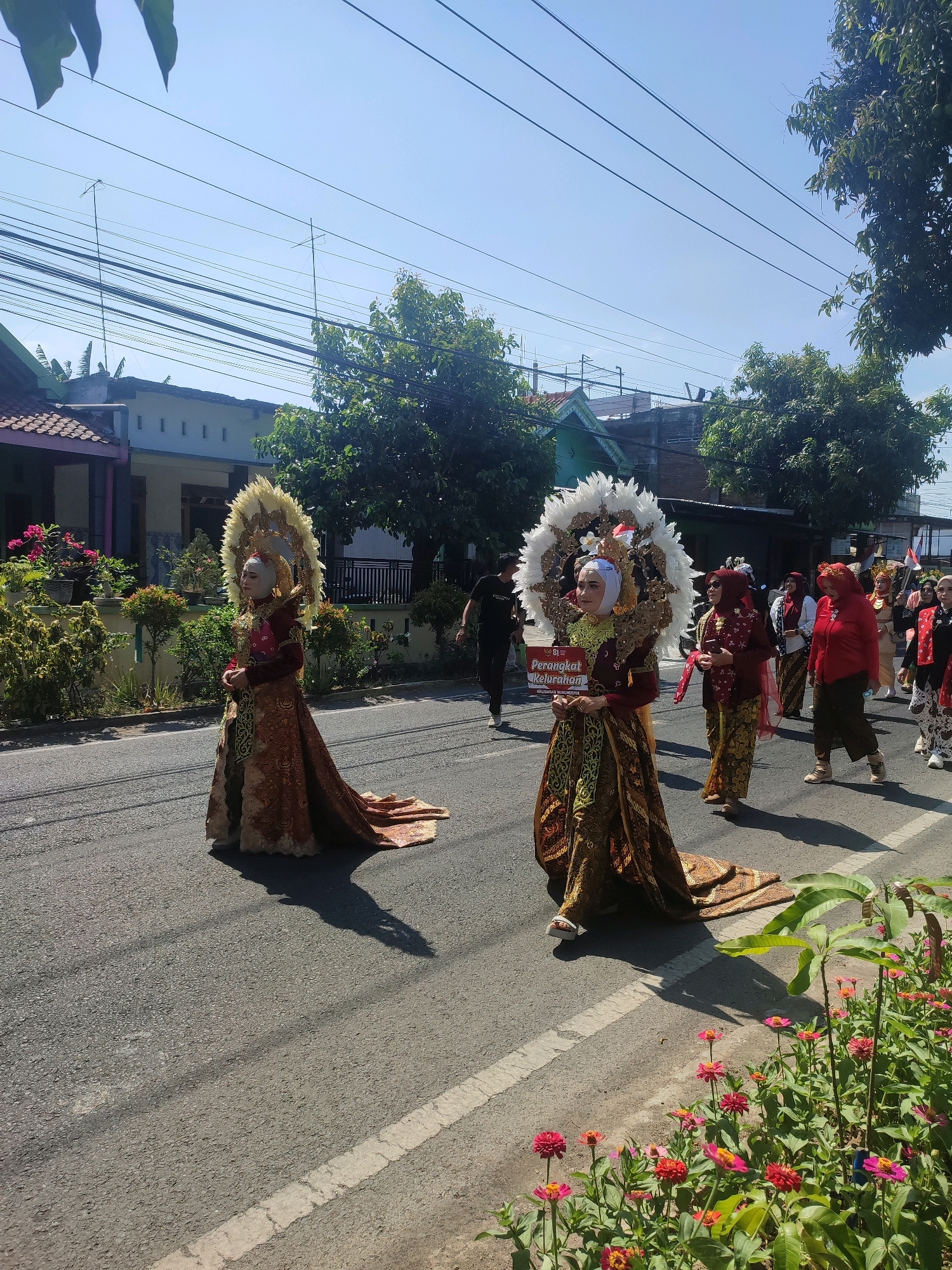 In Nganjuk, Indonesia, two women in elaborate traditional attire participate in a parade. The woman in the foreground is dressed in a dark red and gold patterned garment with a large, feathered headdress. She holds a sign that reads "Perangkat Kelurahan" and "Kepala Lingkungan". Behind her, another woman in a similar outfit walks alongside her. A group of other women, also in traditional clothing, follows them. The parade takes place on a paved road under a clear blue sky, with houses and trees lining the street. The ground is asphalt with a white painted line indicating a lane. Flowers are visible in the foreground on the right side of the image.