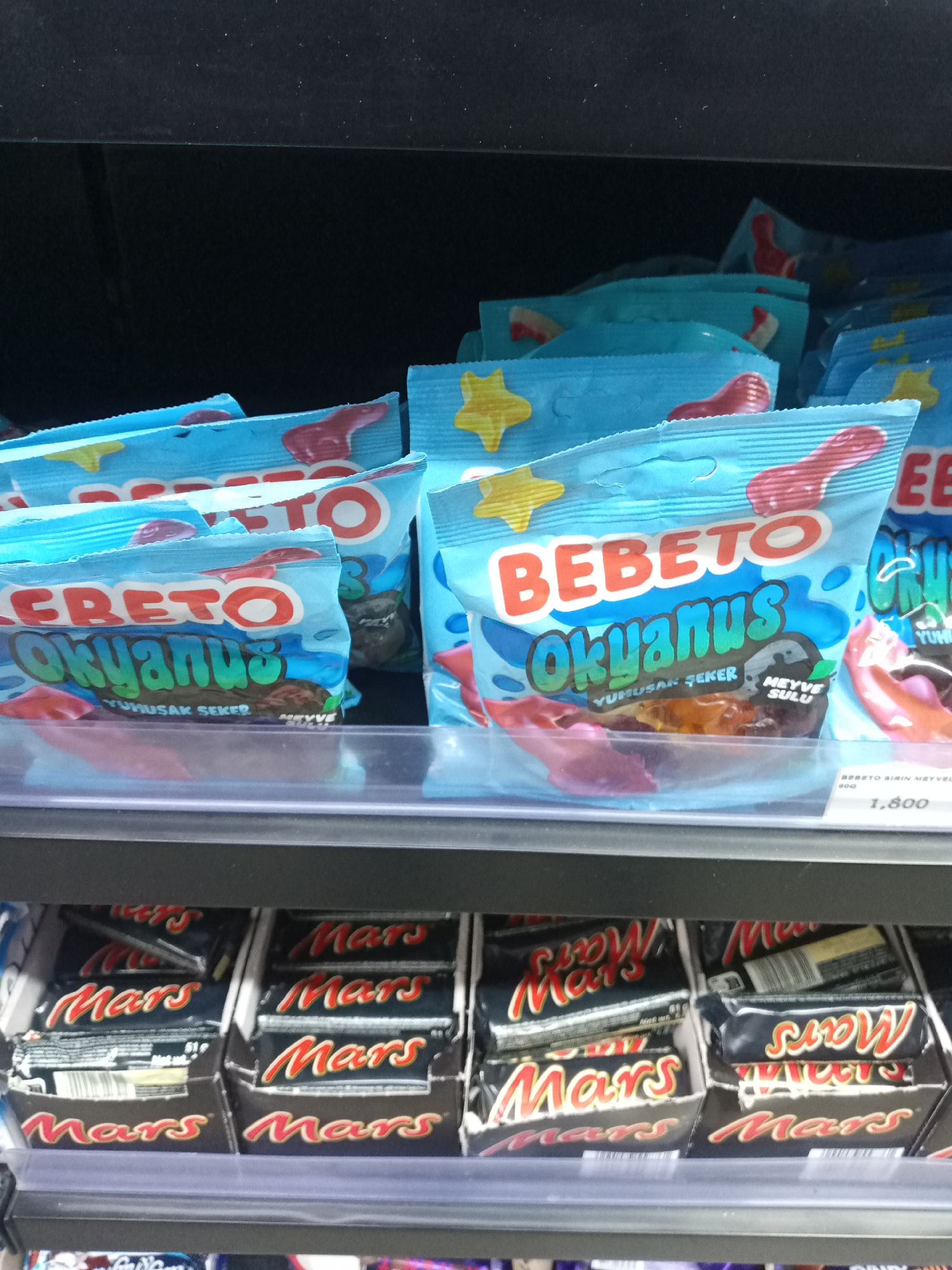A retail store aisle displays two illuminated shelves stocked with packaged confectionery. The top shelf features multiple light blue bags of "BEBETO Okyanus" soft candies. The packaging prominently displays "BEBETO" in red and white lettering, with "Okyanus" in turquoise. Turkish text, "YUMUSAK ŞEKER" (soft candy) and "MEYVE SULU" (fruit juice), is visible on the bags, which also show red gummy shapes and yellow star motifs. A white price tag on the right side of the shelf displays "1,800." The bottom shelf contains numerous individual "Mars" chocolate bars, packaged in black wrappers with the "Mars" logo in red and gold, arranged in cardboard display trays. No people are visible in the frame. The setting appears to be an interior commercial space, likely a supermarket or convenience store. There are no visual cues within the image to confirm the location as Kawaji, Nigeria; the Turkish language on the candy packaging suggests a different geographical context.