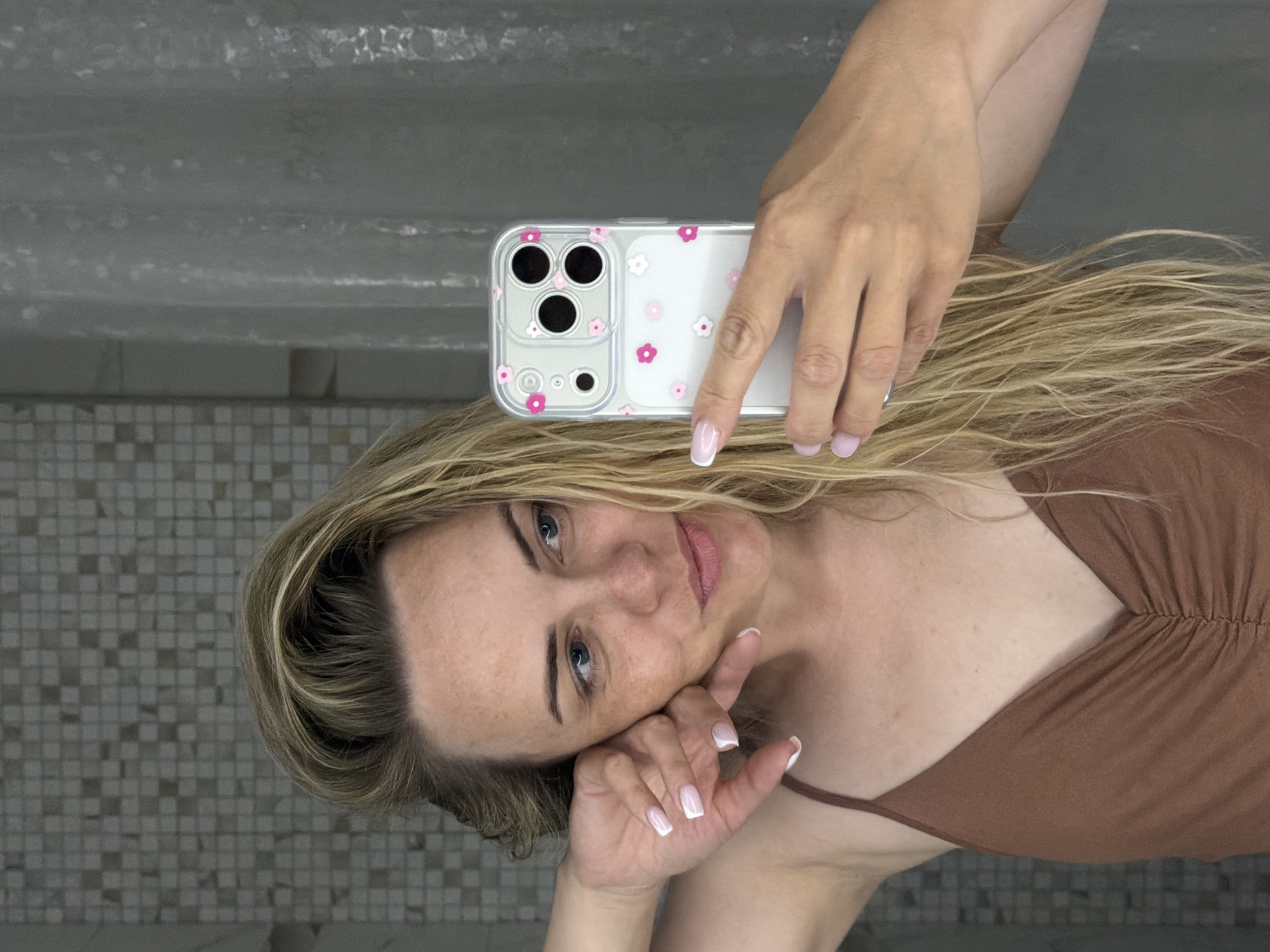 A woman is holding a cell phone above her head, taking a selfie in a bathroom. She has blonde hair, blue eyes, and is wearing a brown top. The phone has a clear case with pink and white flower designs. The background shows bathroom walls with a mosaic tile pattern on the lower half and a plain grey surface on the upper half, suggesting she is likely inside of a residential bathroom. There is no information to suggest the image was taken in Votkinsk, Russia.