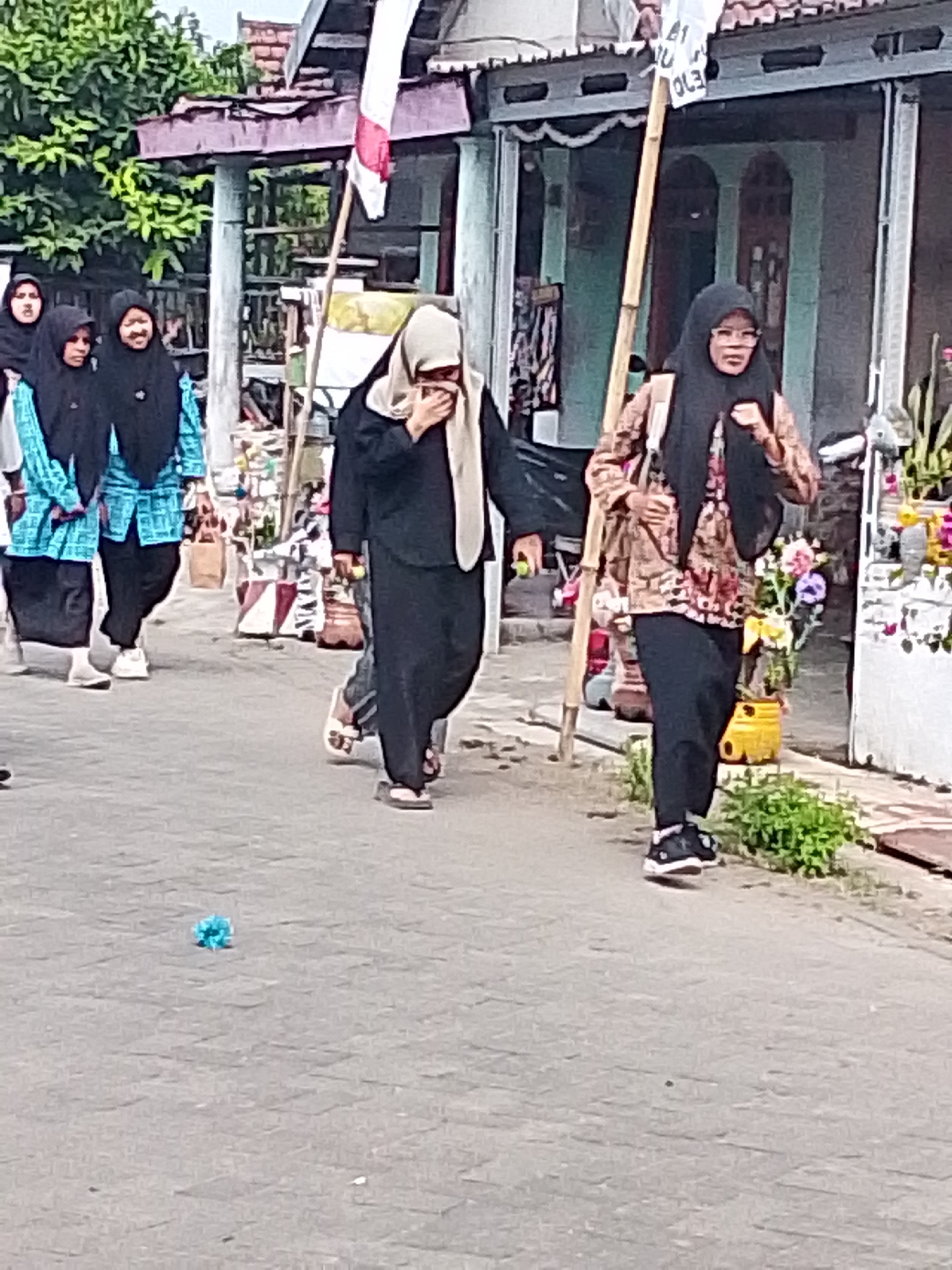 The image depicts a street scene in Bulurejo, Indonesia, with several individuals walking. In the foreground, a person wearing a black headscarf and a patterned brown jacket walks on the right side of the street. They are wearing black pants and black and white sneakers. To the left, two individuals in teal patterned shirts and black headscarves are walking side-by-side, followed by a third person in a black headscarf. Another individual in a black headscarf is walking further to the left. A blue object, possibly a crumpled piece of paper or a small toy, lies on the paved ground in the lower left quadrant of the image. Buildings with visible roofing and structures are present in the background, with some greenery and potted plants also visible. A bamboo pole, likely supporting a flag or banner, is positioned vertically in the center of the frame. The scene appears to be outdoors during the daytime.