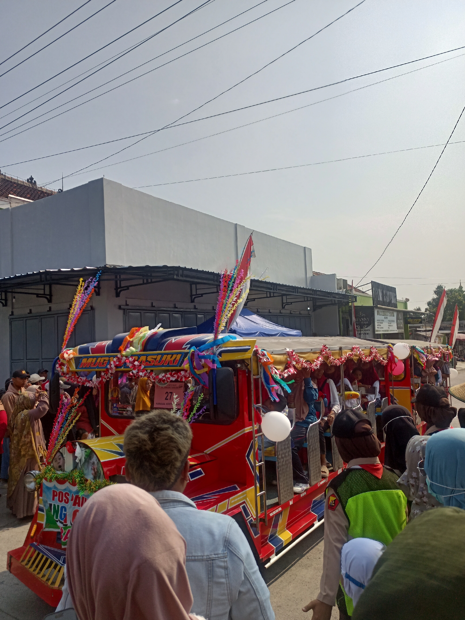 In Gemolong, Indonesia, a vibrant street scene unfolds with a brightly decorated red and yellow vehicle as the central focus. The vehicle, possibly a modified open-air truck or jeep, is adorned with colorful streamers, flowers, and banners, suggesting a festive occasion or parade. Several people are visible, both inside and around the vehicle. Some are seated on the vehicle's structure, while others stand on the roadside, observing or participating in the event. Many individuals are wearing headscarves or hats, typical attire in the region. The backdrop includes commercial buildings with signage and a clear sky, indicating an outdoor daytime setting. The overall atmosphere appears lively and communal, with a sense of celebration.