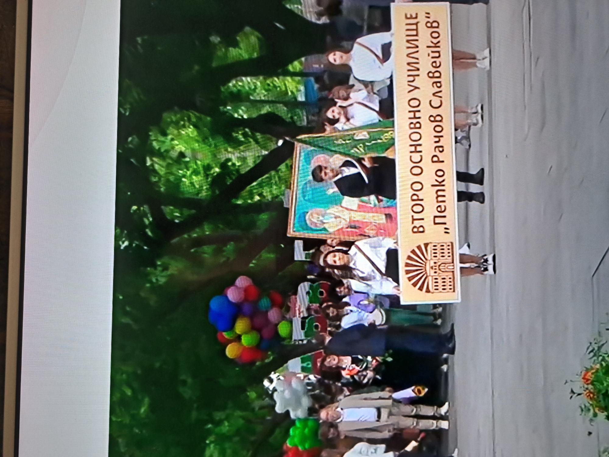A crowd gathers in what appears to be a city park, evidenced by the abundance of green foliage and a paved walkway. The focus of the gathering seems to be a parade or procession, suggested by the presence of balloons, a banner bearing the text "ВТОРО ОСНОВНО УЧИЛИЩЕ „Петко Рачов Славейков“" and a school icon, and an icon. Some individuals in the crowd are wearing formal attire.