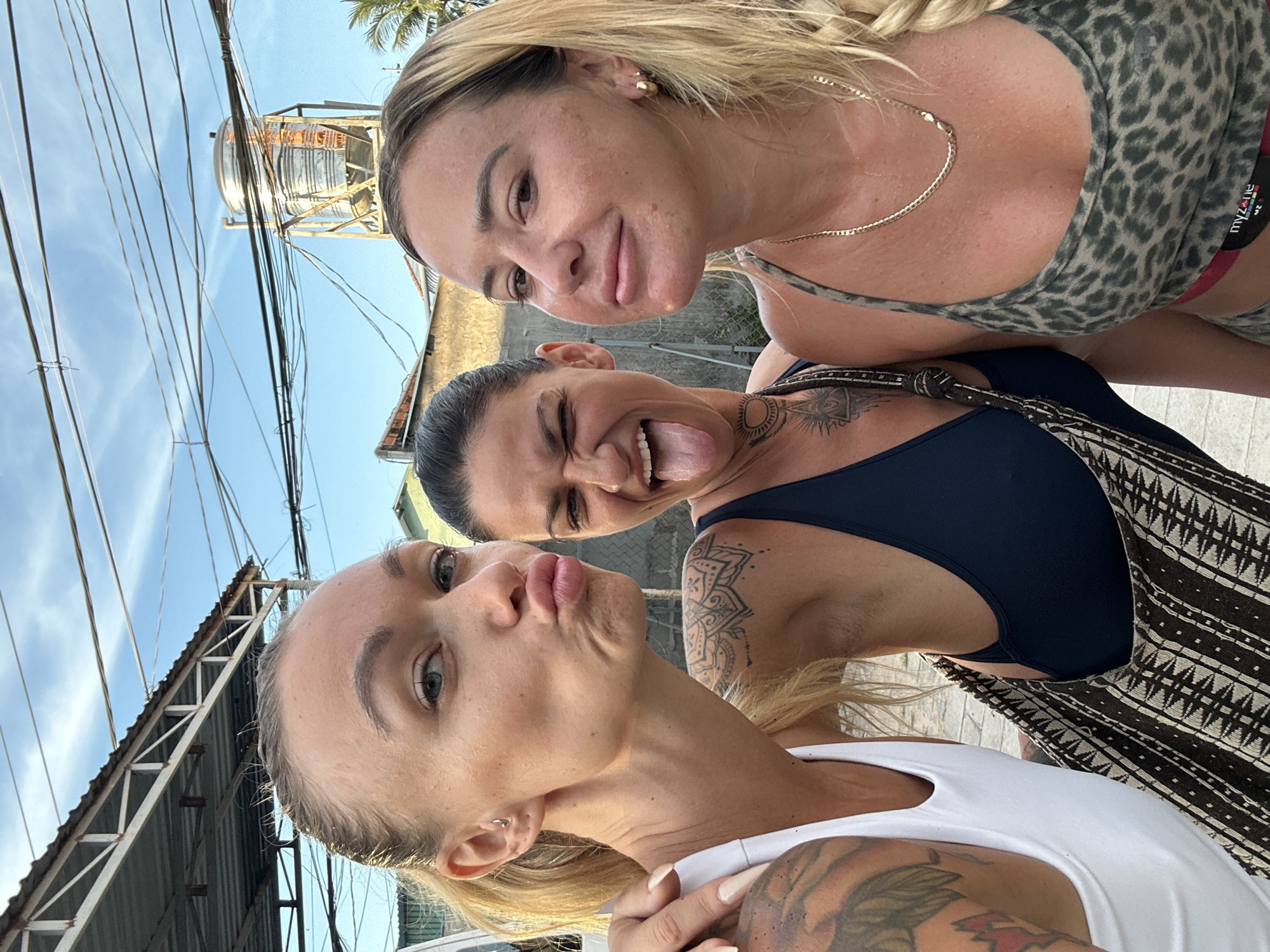 Three women are taking a selfie outdoors. The woman on the left is wearing a white tank top and has her hair pulled back. She is puckering her lips. The woman in the middle is wearing a dark blue sports bra and is sticking her tongue out and winking. She has tattoos on her neck and arm. The woman on the right is wearing a leopard print sports bra and a gold chain necklace. She has blonde hair in a braid. They are standing in front of buildings and a water tank, with power lines overhead. This scene is consistent with the urban and coastal environment found in Mũi Né, Vietnam, known for its beaches and tourist amenities.