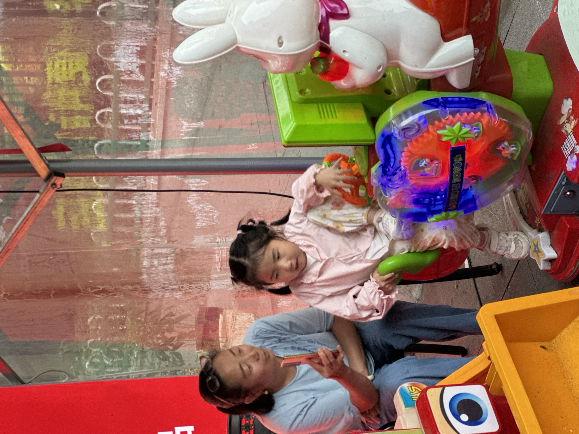 The image shows two people, an adult and a young child, interacting with a coin-operated amusement ride. The adult, wearing a light blue t-shirt and blue jeans, is seated and holding a red object, possibly a phone or toy, up to the child. The child, dressed in a pink outfit, is seated on the amusement ride, which features a white rabbit head design and colorful mechanical components. The child is holding a green object and appears engaged with the ride. The amusement ride is brightly colored with green and red elements, and has a transparent section revealing spinning gears that are illuminated with blue and orange light. A yellow plastic bin is visible to the right, possibly containing sand or other play material. The background appears to be an indoor or covered outdoor space with reflective surfaces showing abstract patterns and colors. Visible text on the ride includes "FASHION TOY" in stylized lettering. The overall scene suggests a playful and casual atmosphere, possibly a public entertainment area or arcade in Chongqing High-tech Zone, China. The lighting indicates it could be daytime, though the exact time of day or weather conditions are not clearly discernible.