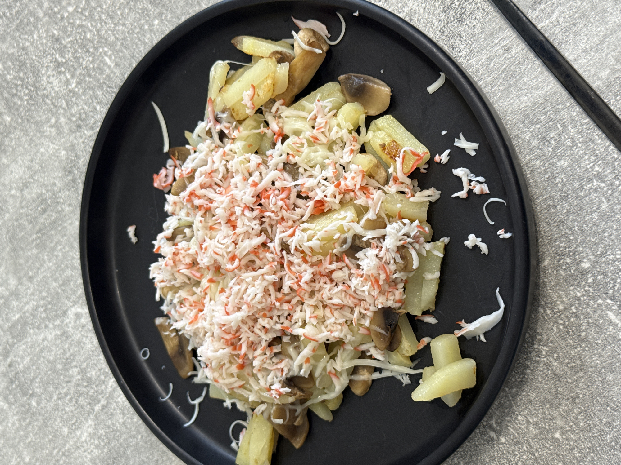A black plate of food is centered on a gray, textured surface. The plate contains what appears to be fried potato wedges, sliced mushrooms, and a generous topping of shredded imitation crab meat. The food is arranged in a mound, with some pieces spilling over the side of the plate and onto the surface. The lighting suggests an indoor setting, possibly a kitchen or dining area. There are no people visible in the image. The gray surface appears to be a countertop or tabletop with a speckled pattern.