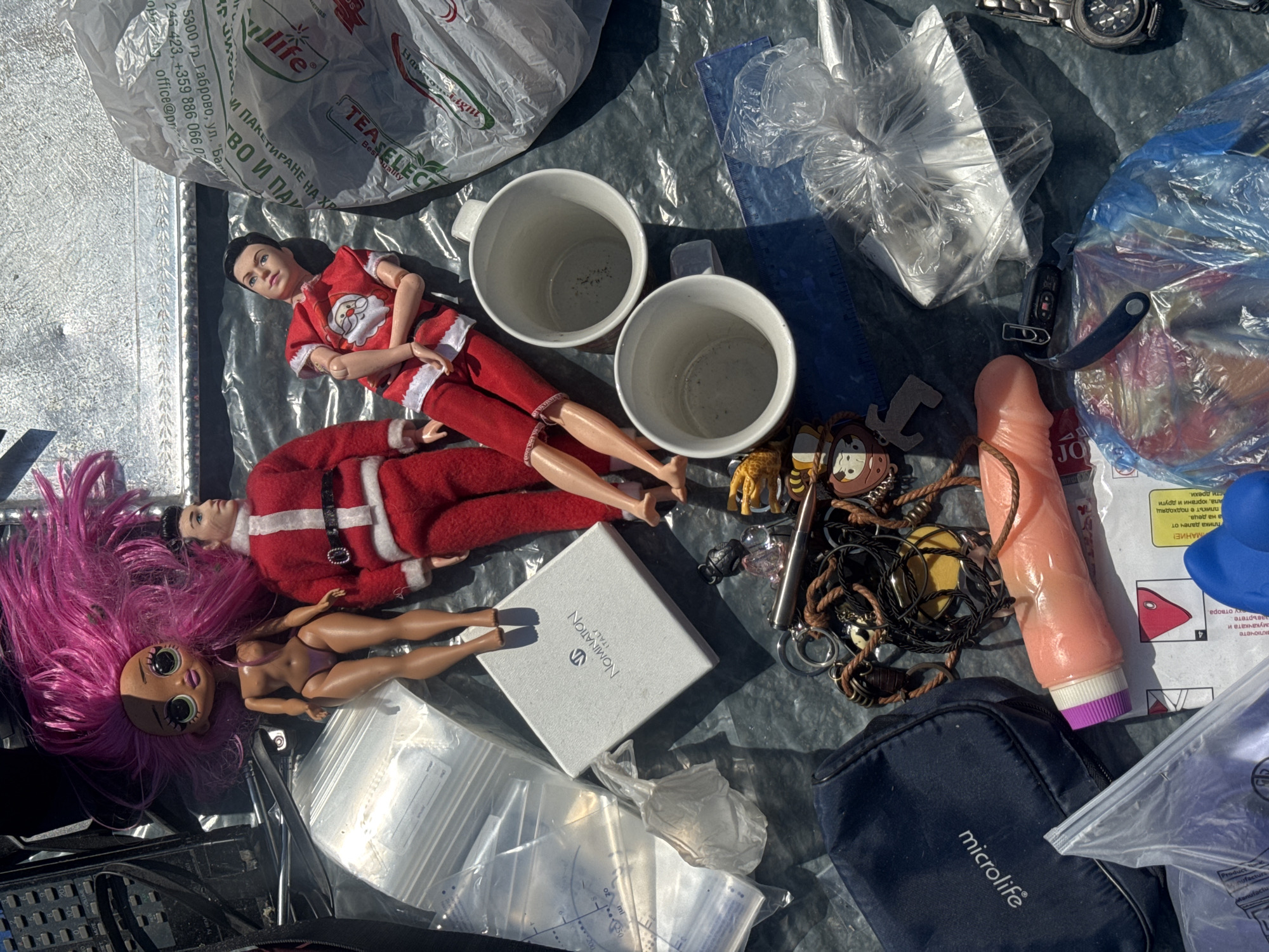 An overhead, high-angle shot captures a collection of items laid out on a dark, possibly tarpaulin surface, suggesting an outdoor setting, perhaps a flea market or informal selling space in Sofia, Bulgaria. The lighting indicates daytime, with strong shadows cast by the objects.
Dominating the upper portion are two dolls: one with vibrant pink hair and a tan complexion, partially clothed in a purple bikini-like garment, and another male doll dressed in a red and white Santa Claus-themed outfit. Next to the dolls are two white ceramic mugs. To the left, a white jewelry box with "NOMINATION ITALY" printed on it rests near clear plastic bags containing various items.
Below these, a dark blue pouch with "microlife" printed in white is visible. A pink, phallic-shaped object with purple and white detailing lies horizontally across the lower-middle section of the image, partially obscuring a small instruction booklet with Cyrillic text. A collection of keychains, including charms with cartoonish faces and metallic elements, are scattered. Additional objects include a metallic pen-like item, a blue ruler, and what appears to be a disassembled electronic device or a power bank.
In the lower right, a metal container wrapped in plastic and a wristwatch with a black strap are present. The overall impression is of miscellaneous, possibly second-hand items arranged for display or sale. The background is blurred but appears to be a textured surface.