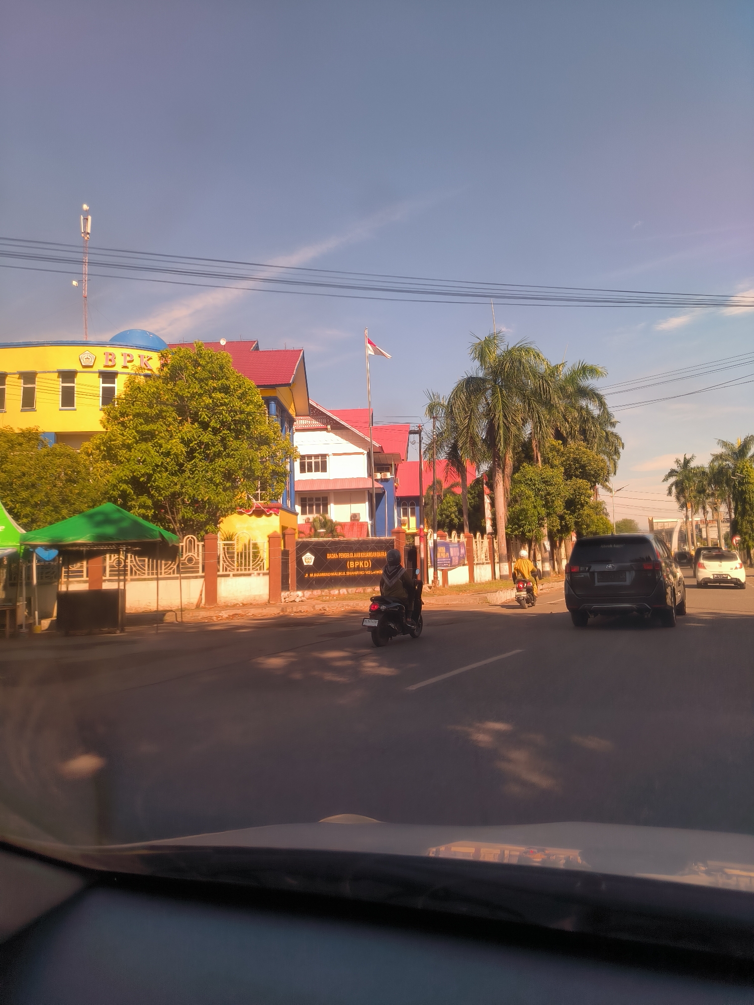 The image captures a street scene in Lhokseumawe, Indonesia, viewed from inside a vehicle, with the dashboard and windshield reflections visible in the foreground. The time of day appears to be late afternoon or early evening, characterized by warm, golden sunlight casting long shadows and a clear blue sky with faint wisps of clouds.
On the left, a prominent multi-story building stands out with a bright yellow facade, a blue dome on its roof, and red-tiled sections. Large green trees partially obscure its lower levels. Visible text on the building identifies it as "BPK," and a sign below reads "BADAN PENGELOLAAN KEUANGAN DAERA (BPKD)." Another line of text is partially visible as "MUHAMMAD MALIKUL SHAHR IND IS LOOKSI" (from OCR). Adjacent to this building are tall palm trees and another structure with a white facade and red roof. A red and white flag, likely the flag of Indonesia, flies from a flagpole near the buildings.
In the midground, a paved road carries several vehicles, including a dark SUV and a white car in the distance. Two motorbikes are also visible, one with a rider wearing a brown jacket and another with a yellow-clad rider. Green canopy tents are set up on the sidewalk near the yellow building, and a decorative fence runs along the property line. The scene depicts a typical urban environment with daily traffic and government buildings.