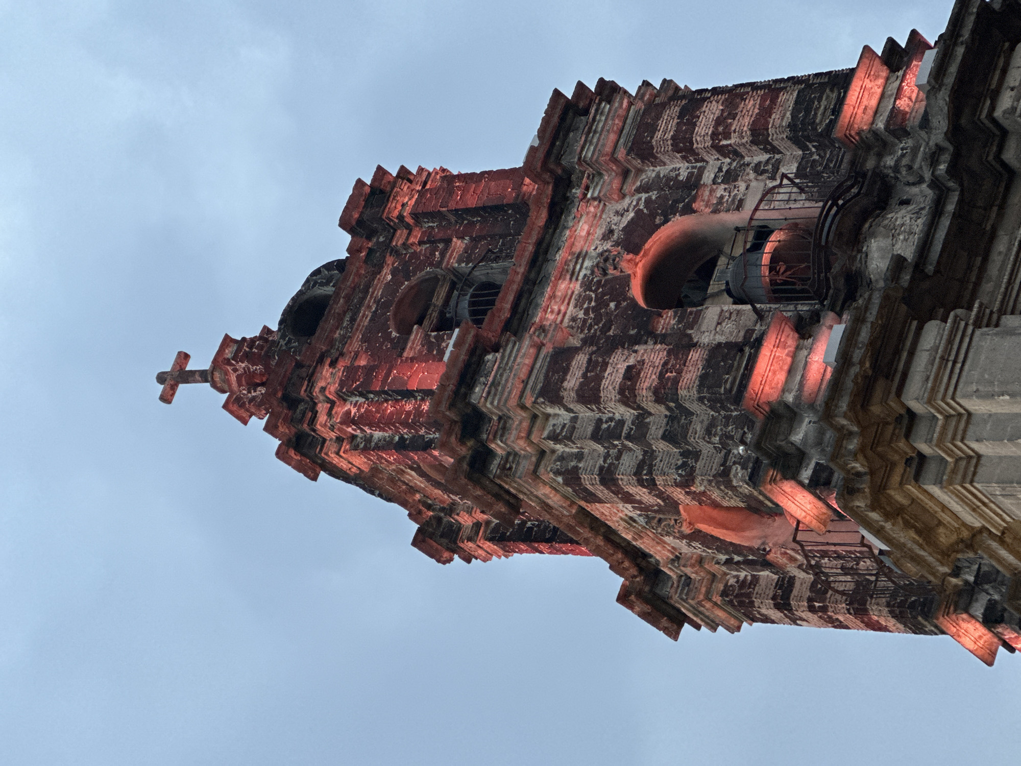 Low-angle view of the side of a red brick church tower, taken in Mexico City, Mexico. The tower has a zigzag pattern with some red lighting. There is a cross on top of the tower, and the sky is in the background.