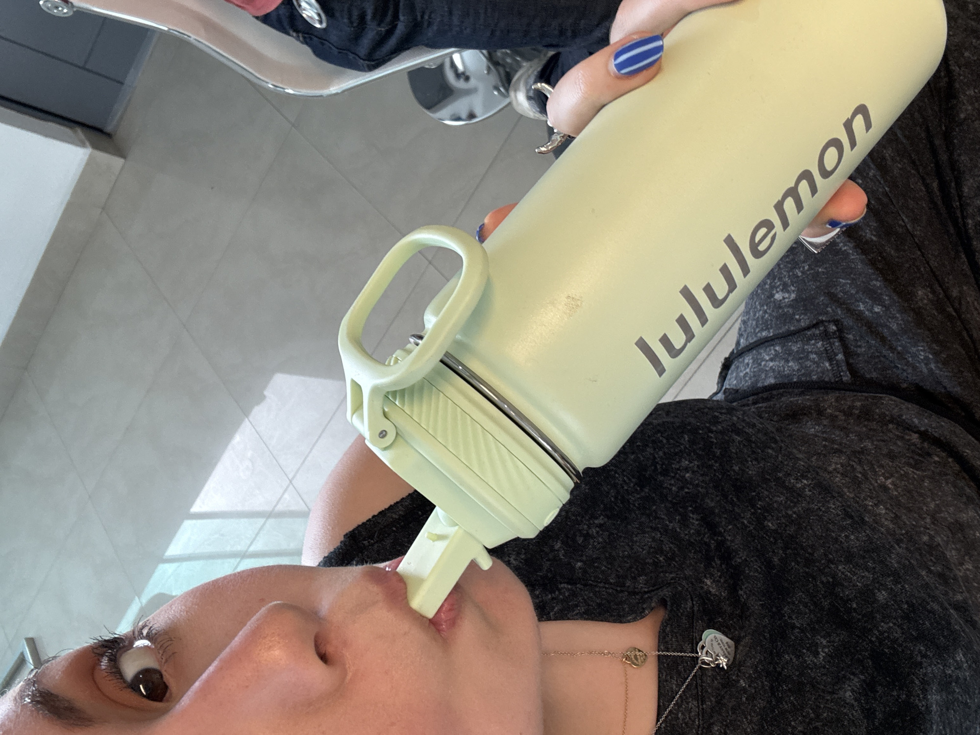 A person is drinking from a light green "lululemon" water bottle. The person is wearing a dark-colored top and has blue and white striped nail polish. A silver necklace with a charm is visible. The background shows a tiled floor and part of a chair. I cannot identify the location within Santa Clara Ocoyucan, Mexico, or if this image was taken there.
