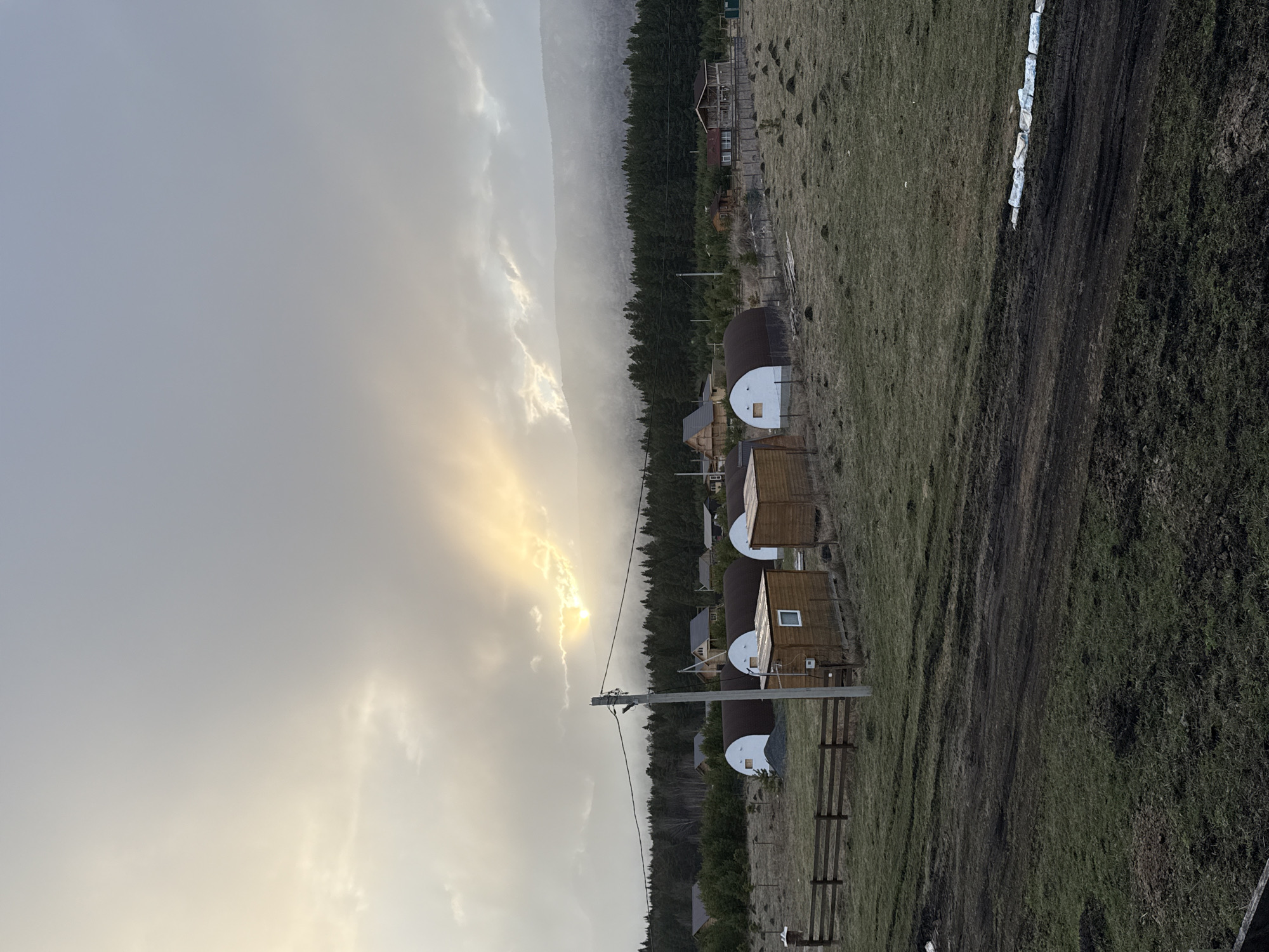 The image shows a landscape in Tyulyuk, Russia, at what appears to be dusk or dawn. The sky is overcast with clouds, with the sun partially visible. A line of small, cylindrical cabins with wooden exteriors and white domed roofs is situated along the edge of a grassy field. Adjacent to the cabins is a dirt road. A forested area is visible beyond the cabins. A set of power lines runs parallel to the cabins. In the distance, a hazy mountain range can be seen. The overall scene evokes a remote, rural setting.