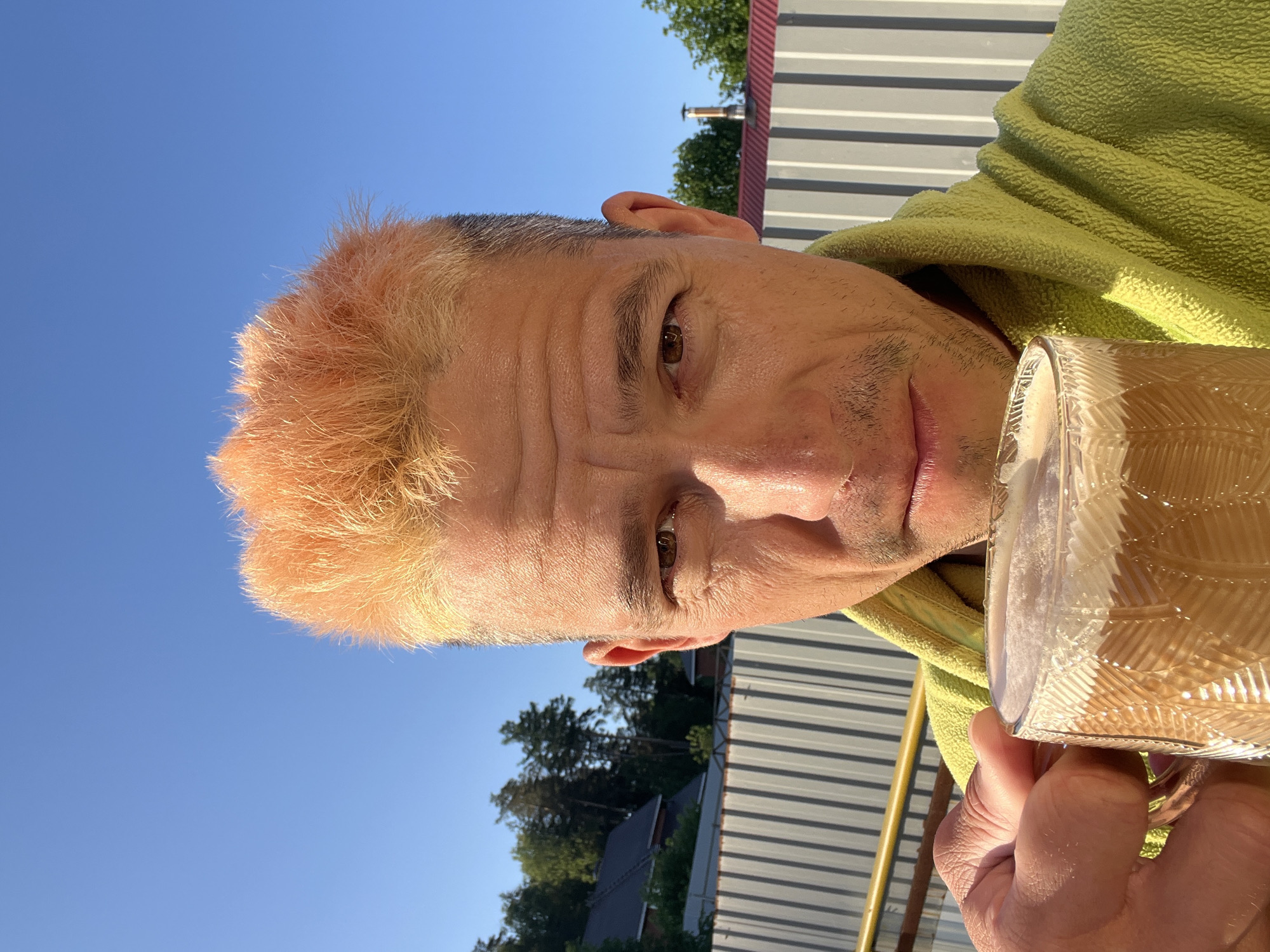 A middle-aged man with peach-colored hair is holding a glass of light-colored liquid, likely a drink, in his right hand. He is wearing a light green fleece jacket. The man is in an outdoor setting with a view of a corrugated metal building, trees, and a blue sky. Based on the presence of what appears to be typical Russian suburban housing and foliage, it's likely this photo was taken in a suburban area. Given the information provided, it is probable that this scene is located in a suburban part of the city of Chaikovsky, Russia. The photo appears to be a selfie taken from a low angle.