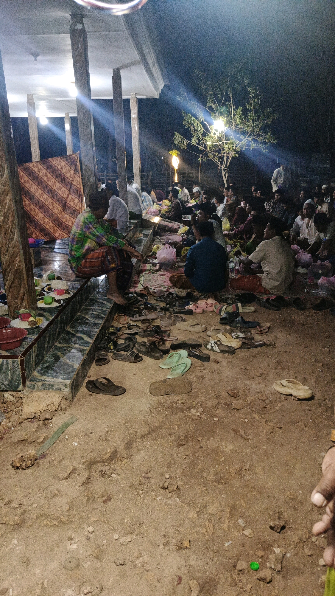 In Sokaramme Timur, Indonesia, a group of people are gathered outdoors at night, likely for a communal meal or religious event. The scene is illuminated by artificial lights, casting long shadows. Many individuals are seated on the ground or on a raised tiled platform, some facing each other. Food is visible on plates and in bowls, spread out on mats and the ground. A variety of footwear, including sandals and flip-flops, are scattered around the seating area. In the background, trees and a structure with pillars are discernible. The ground appears to be dirt and gravel.