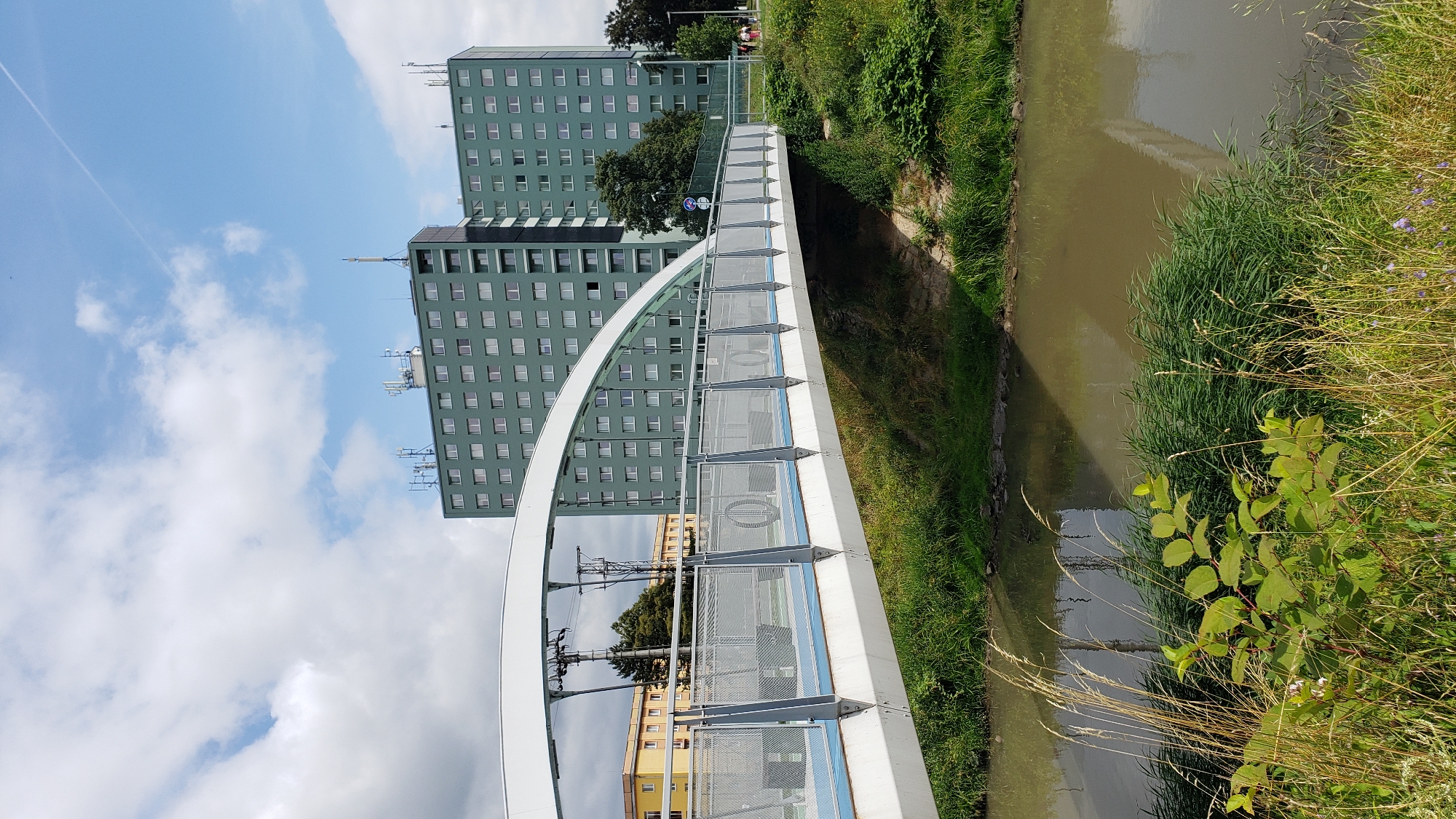 This is a photograph of a pedestrian bridge arching over a moderately sized body of water, likely a river or canal. The bridge is modern in design, featuring a white curved superstructure and a deck with metal railings and what appears to be a blue surface. Beyond the bridge, two large, multi-story residential buildings dominate the background. These buildings are identical in design, painted a muted green, and feature numerous rectangular windows. Antennae and communication equipment are visible on the rooftops of these buildings. In the middle ground, a yellow building with multiple floors is visible to the left of the green buildings. The scene is set in an urban environment, with lush green vegetation, including tall grasses and bushes, lining the banks of the water. The sky is partly cloudy with blue patches and a visible contrail from an aircraft. A few people are walking on a path to the right of the green buildings, indicating public accessibility to the area. The location is identified as being in the city of Prešov, Slovakia, specifically the Sídlisko Sekčov residential area, near the main road.