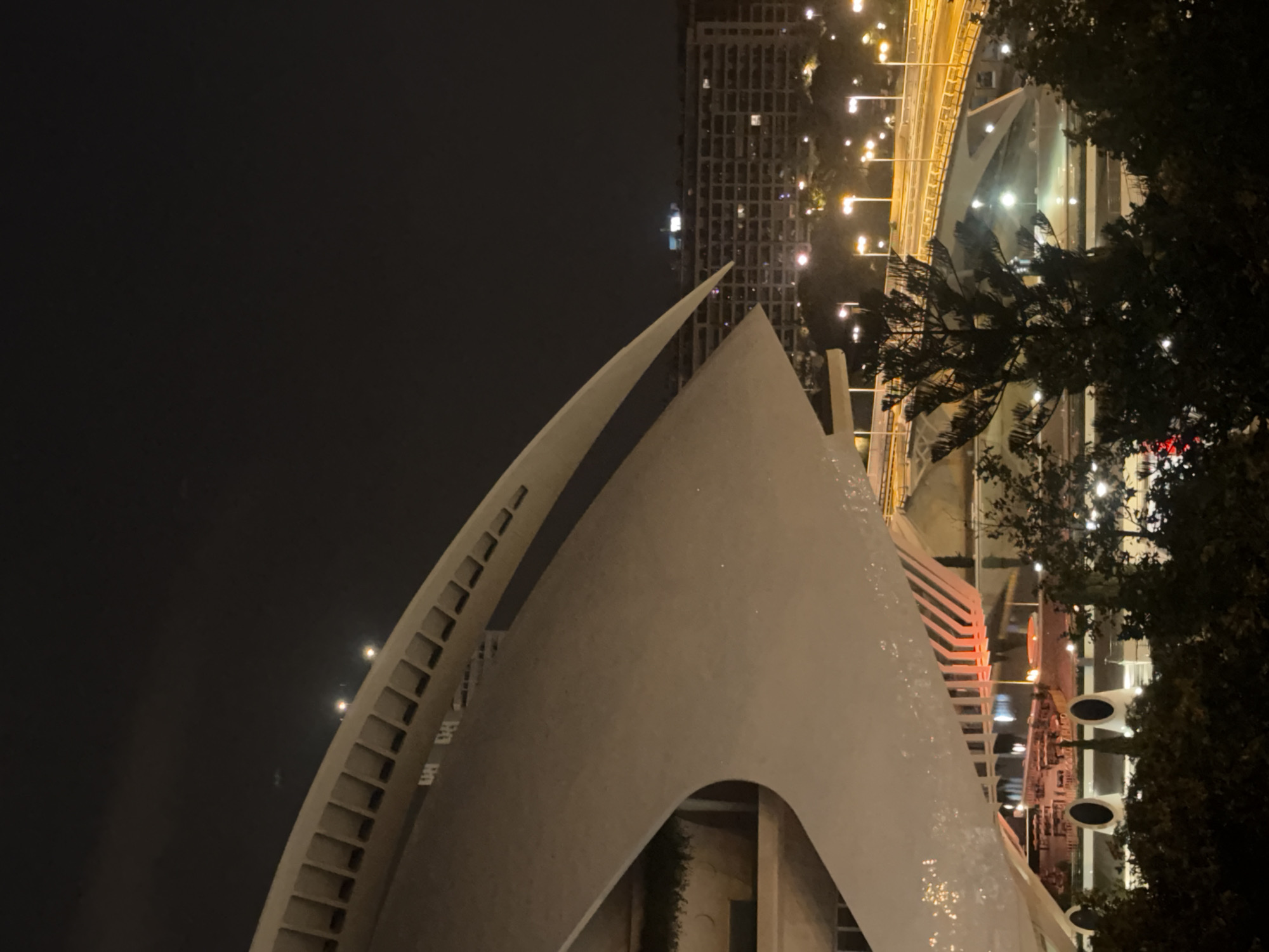 The image captures a nighttime view of the Hemisfèric building in Valencia, Spain. The architecture is modern and futuristic, with a large, white, curved structure dominating the foreground, its smooth surface reflecting the ambient light. In the background, a multi-lane highway illuminated by streetlights stretches across the scene, with vehicles visible on the road. Beyond the highway, a large residential building with numerous lit windows is visible, indicating a densely populated urban area. The sky is dark, suggesting it is nighttime. The foreground also features a section of the Hemisfèric’s exterior with a rhythmic pattern of indentations, and below it, what appears to be an outdoor seating area with tables and chairs, illuminated by warm red and orange lights. Trees and dark foliage are present in the lower portion of the image, framing the view of the illuminated urban landscape.