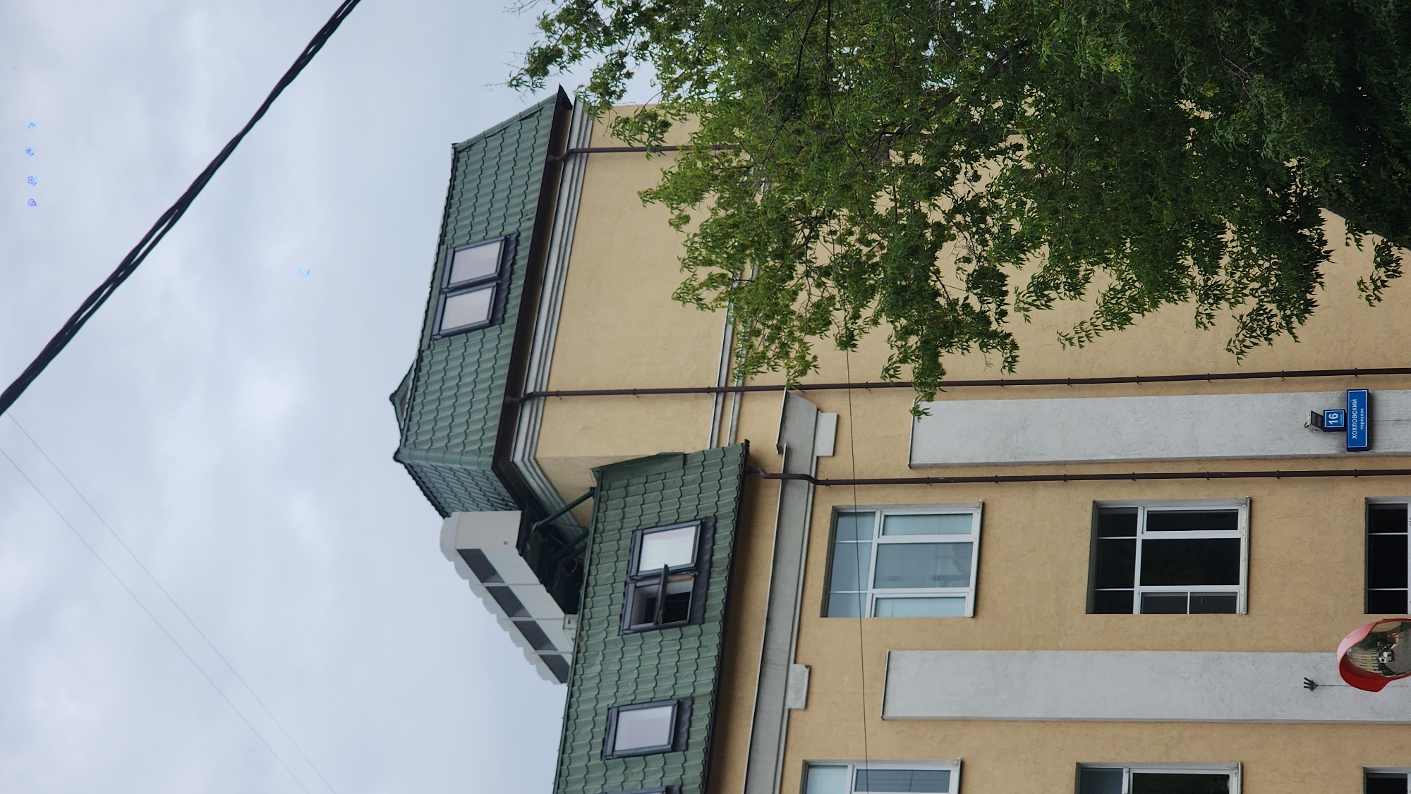 This media file shows a section of a multi-story building with a light yellow facade and a green, corrugated roof. The building has several windows, some with white frames and others with darker frames. There are also some horizontal decorative bands on the facade. A tree with green leaves is partially visible in the upper right corner, casting shadows on the building. Electrical wires are visible in the upper left and extending diagonally across the frame.
A blue street sign with white lettering and numbers is affixed to the building, displaying "16" and Russian text that appears to read "ХОЛМОГОРСКИЙ переулок" (Kholmogorsky Lane). In the lower right, a red and chrome convex mirror is attached to the building, reflecting a distorted image of the surroundings. Some subtle blueish, lens flare-like artifacts are present in the sky. The sky is overcast and appears to be a pale gray, suggesting cloudy weather. The image is taken from a low angle, looking upwards towards the building and sky. There are no people or discernible activity taking place. The time of day is unclear due to the overcast conditions, but the lighting suggests it is daytime.