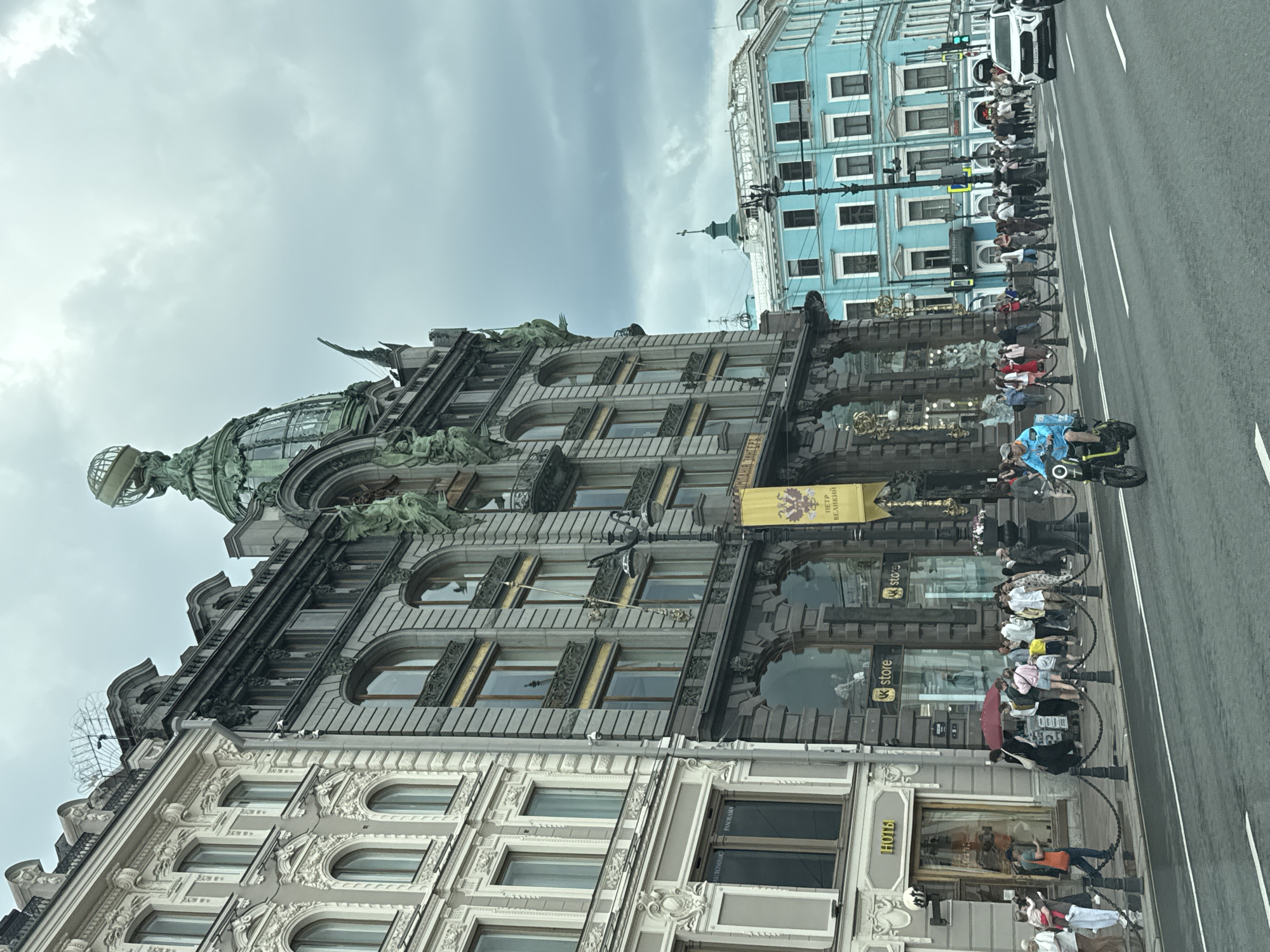 This image depicts a street scene in Saint Petersburg, Russia, likely near Nevsky Prospekt, a major thoroughfare. The dominant structure is the Singer House, an iconic Art Nouveau building with a distinctive glass dome topped by a globe supported by an eagle. The facade of the Singer House features ornate architectural details, including sculptures and large arched windows. To the left of the Singer House, an adjacent building displays a pale, decorated facade with arched windows. Across the street, a vibrant turquoise building with numerous windows stands out. The street is paved with asphalt and marked with white lane lines. A diverse group of pedestrians are seen on the sidewalk, some walking, others standing in conversation, and a few are holding umbrellas. One individual is riding a scooter in the foreground, moving from left to right. A white SUV is visible on the right side of the street, indicating vehicular traffic. The sky is overcast with dark clouds, suggesting a cloudy day.