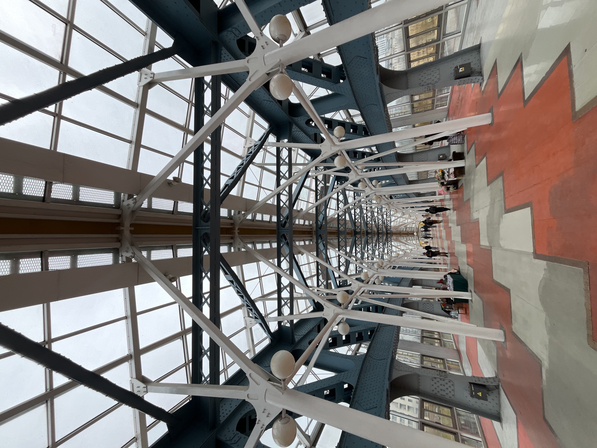 The image shows the pedestrian bridge over the Moskva River in Moscow, Russia, known as the Bogdan Khmelnitsky Bridge. The perspective is from within the enclosed, glass-roofed section of the bridge. Structural steel elements, painted in shades of blue and white, form an intricate lattice overhead. Rounded light fixtures hang from the structure. The walkway is adorned with a red and grey zig-zag pattern. People are walking along the bridge in the distance. The windows to the sides offer views of the city.