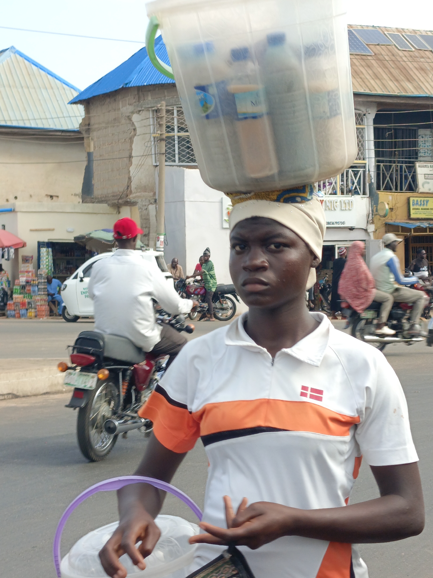 In Akwanga, Nigeria, a person is carrying a large, clear plastic bucket filled with bottled drinks on their head. The person is wearing a white shirt with an orange stripe across the chest and a small Danish flag emblem. They are also holding a smaller, clear plastic bucket with a purple handle in their left hand. The person appears to be standing on a street with motorcycles and pedestrians in the background. The surrounding environment includes buildings with varying architectural styles, some with corrugated metal roofs and others with more modern designs. Solar panels are visible on the roof of one building. There are shops with signage in the background, and overall, the scene depicts a bustling street environment in a city.
