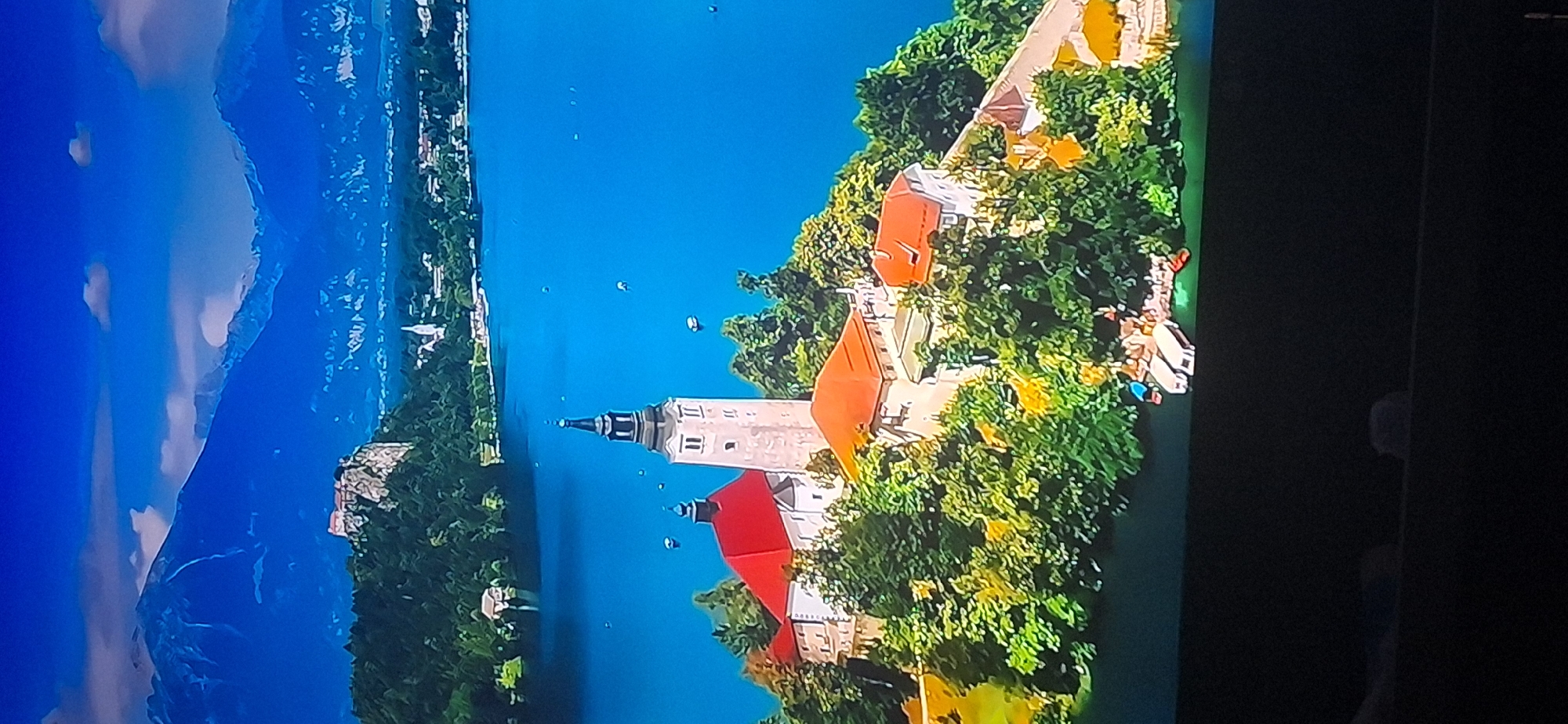 The image depicts a picturesque landscape with a prominent island in a bright blue lake. On the island, a complex of buildings is situated, featuring a tall bell tower with a black spire, a larger building with a distinctive red roof, and several other structures with orange-tiled roofs, all surrounded by lush green trees. In the background, a dramatic mountain range under a blue sky with scattered clouds dominates the horizon. A small castle or fortress is visible on a rocky outcrop to the left of the mountains. Several small boats are dotted across the lake and near the shore of the island. The scene is bathed in bright, natural light, suggesting a clear day. There are no people visible in the image. The overall impression is one of serene natural beauty and historical architecture. This image does not depict the city of Bila Tserkva, Ukraine.