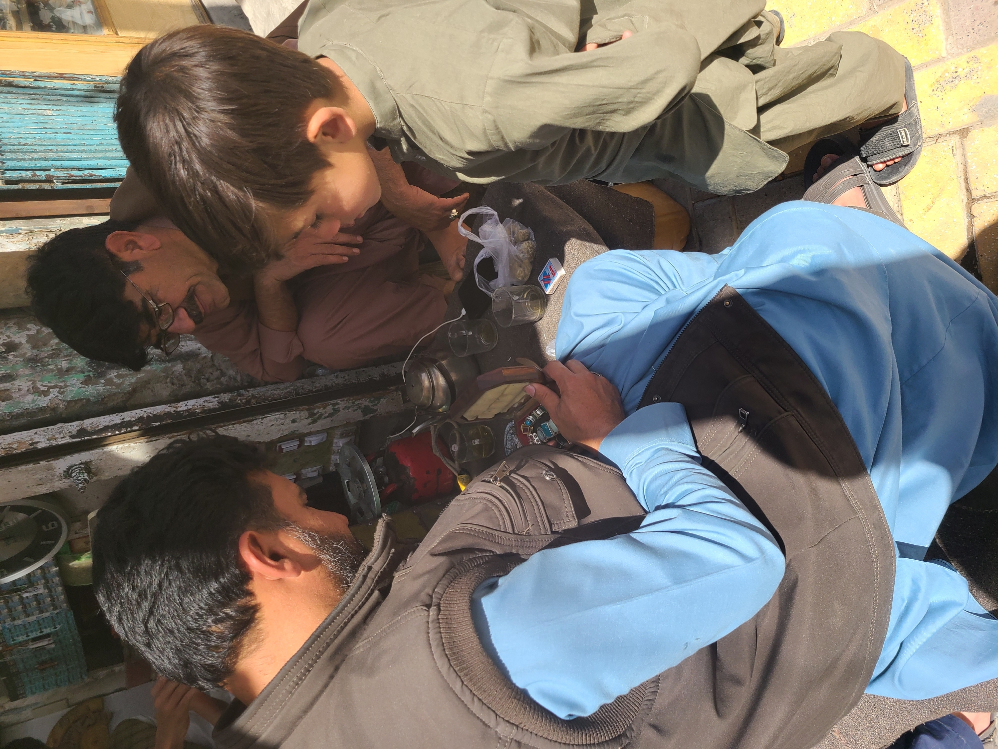 In Quetta City Tehsil, Pakistan, a group of four men are gathered outdoors. One man is wearing a brown shirt and glasses, another is in light green clothing, a third wears a blue top with a dark vest, and the fourth is partially visible. They are seated around a collection of items including glass jars, a plastic bag containing a substance resembling marijuana, and what appears to be a matchbox. The group seems to be involved in preparing or using the contents of the bag. The setting appears to be on a sidewalk or paved area near a building with teal-colored shutters.