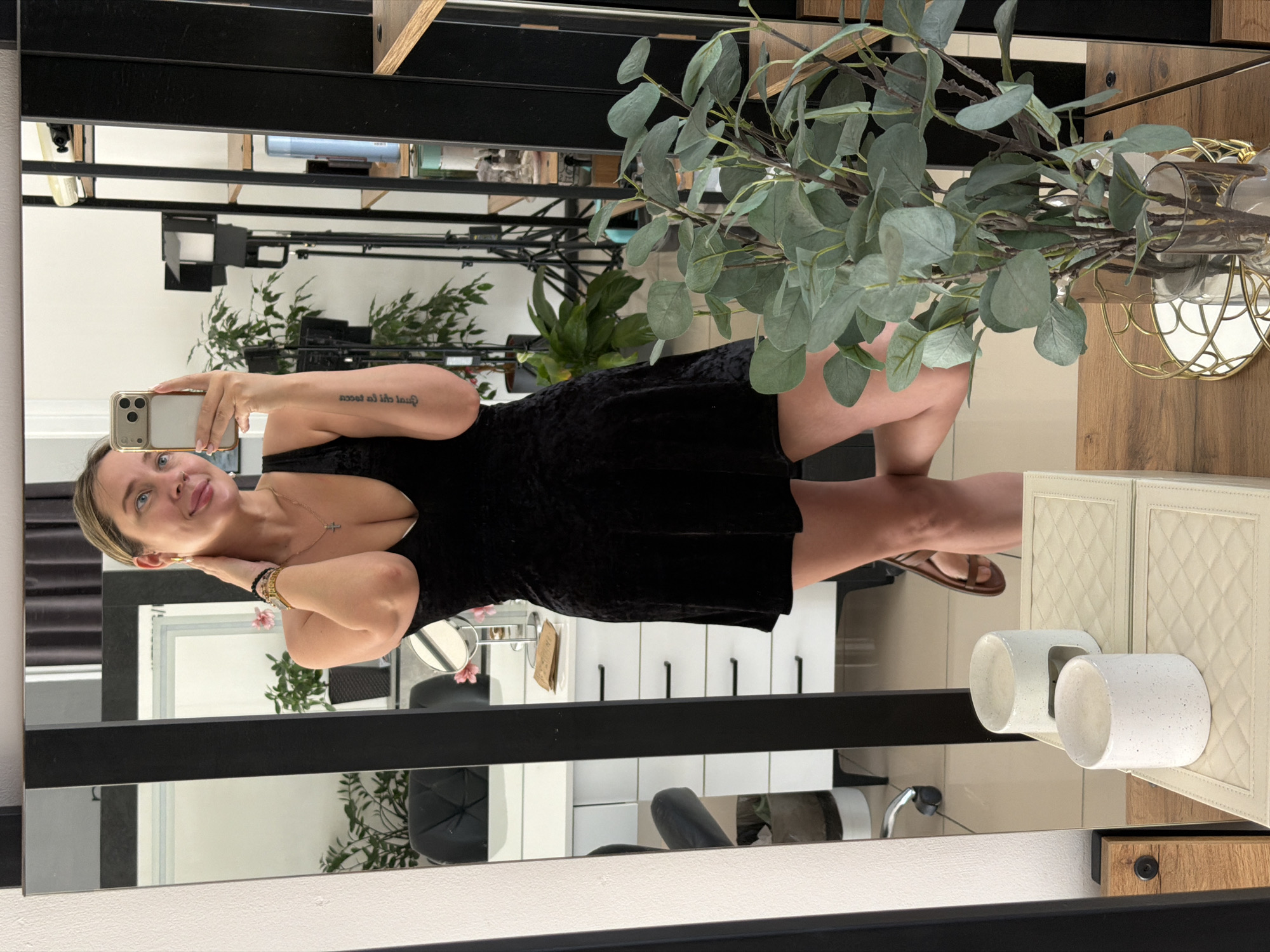 A young woman is captured in a mirror selfie taken indoors in Chaikovsky, Russia. She is smiling, with her head slightly tilted, and her blonde hair pulled back. She wears a black velvet or velour mini-dress, a gold necklace with a cross pendant, and bracelets on her left wrist (one gold, one black beaded). Her right arm, adorned with a tattoo that appears as "sces ed ihte lau" in the mirror (reversing to "love is the answer"), holds a light-colored smartphone with multiple cameras, likely an iPhone, which she uses to take the photo. Her left hand gently touches her neck. She stands with her feet visible, wearing light-colored sandals.
The setting appears to be a well-lit interior, possibly a salon, dressing room, or bathroom, characterized by large mirrored surfaces framed with black and wood-like elements. Reflections reveal various objects and parts of the room. To the left of the woman, reflected elements include white walls, a dark plant in a pot, and a dark chair. To her right, a decorative plant with green, roundish leaves (resembling eucalyptus) is displayed in a clear vase, alongside a gold-colored wireframe light fixture. Further reflections show shelves stocked with bottles and other containers, and more greenery. In the lower right corner, directly visible on a surface in front of the mirror, is a light-colored, possibly cream-quilted box or cabinet, topped with two white cylindrical holders. The overall atmosphere is bright and casual, and the woman appears happy or content.