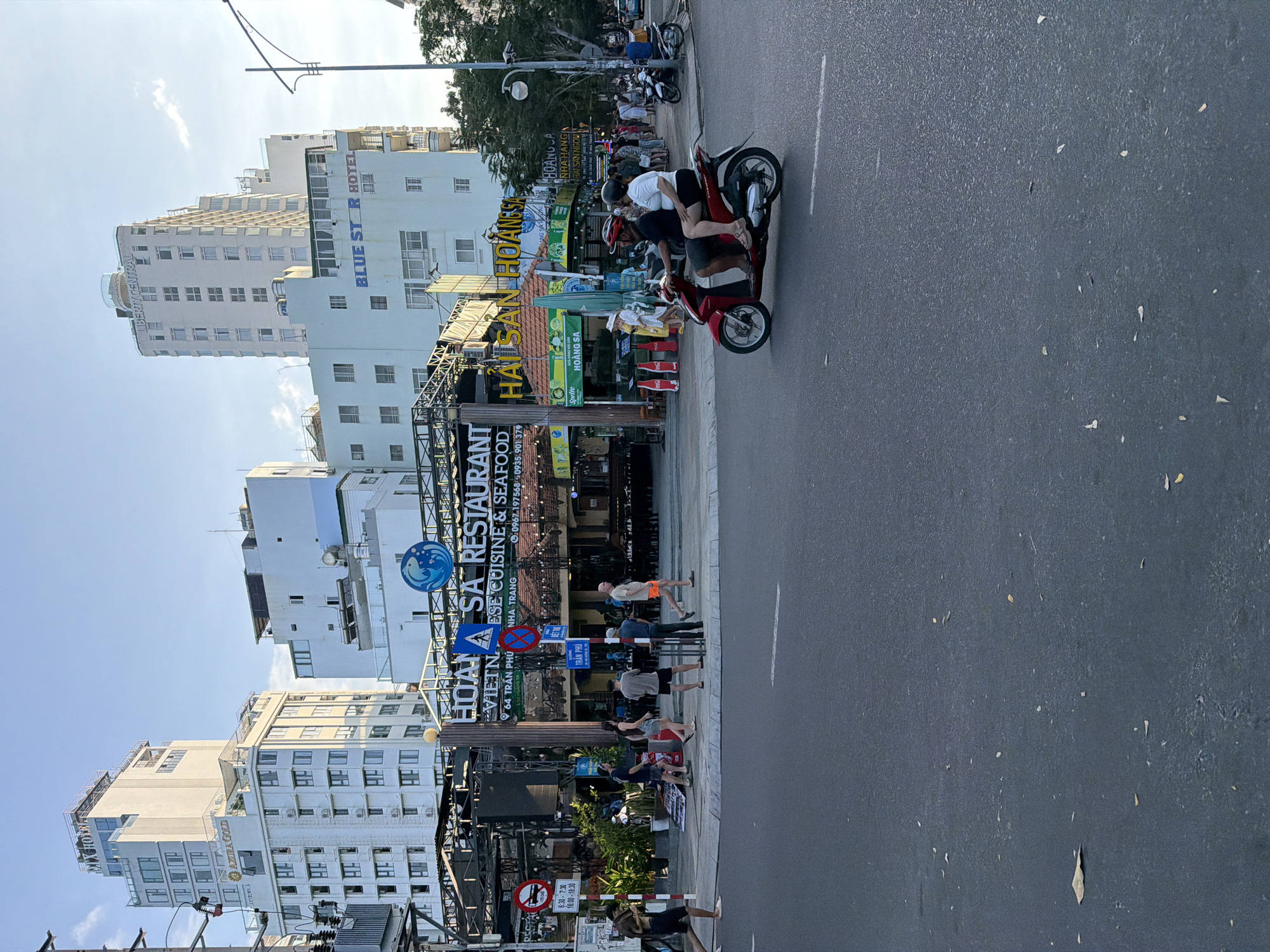 This image captures a street scene in Nha Trang Ward, Vietnam, specifically at 64 Trần Phú Street, near its intersection with Hoàng Sa Street. The central feature is the "HOÀNG SA RESTAURANT," a Vietnamese cuisine and seafood establishment, identified by its large, illuminated sign and visible phone numbers (0967.197.568, 0935.901.379). The restaurant building has an open-air or semi-open dining area with numerous small lights, green foliage, and several large red Coca-Cola shaped bins or decorations. A green Sprite umbrella is also visible near the restaurant's entrance.
In the background, the skyline is dominated by several tall, multi-story buildings, including the "AZURA GOLD" hotel on the left and the "LIBERTY CENTRAL" and "BLUE STAR HOTEL" on the right, all appearing modern and light-colored.
The scene takes place during what appears to be late afternoon or early evening, with a clear to lightly clouded sky and soft, ambient lighting. The asphalt road in the foreground is clean, featuring a white dashed line marking a lane and scattered small debris, such as leaves.
On the road, a red scooter carrying two individuals is in motion, traveling from right to left. The driver wears a red helmet and dark clothing, while the passenger is dressed in a white t-shirt, dark shorts, and a dark helmet. Several other motorbikes are parked further down the street on the right.
The sidewalk on the left is populated by several pedestrians. A person in dark shorts and a t-shirt walks while seemingly looking at a mobile phone. A group of approximately five individuals, casually dressed, walks along the sidewalk. More people are gathered near the restaurant's entrance.
Traffic signs are visible on the left, including a "No Stopping" sign with time restrictions (8:30-7:30 and 16:00-18:00), another circular red prohibition sign, and a blue directional sign indicating "TRẦN PHÚ" to the left and "HOÀNG SA" to the right. A utility pole with transformers and overhead wires is present on the far left of the frame.