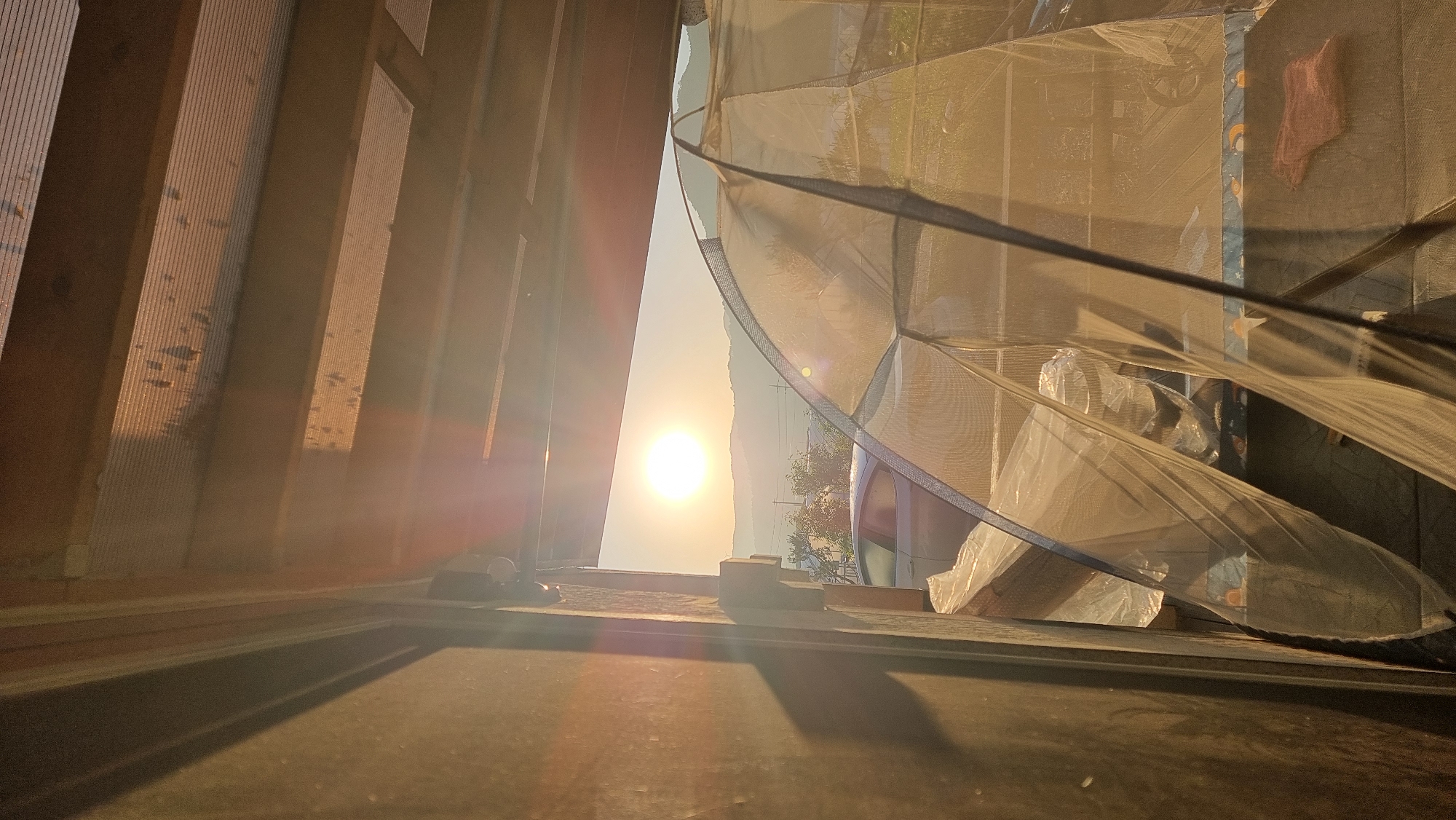 The image, likely taken in Dunnae-myeon, South Korea, shows a low-angle view of a structure with sunlight streaming through an opening. To the left, there's a portion of a wooden structure, possibly a fence or building framing, with vertical planks casting shadows. In the center, a bright light source, presumably the sun, is visible through the opening, creating a strong glare. To the right, a draped, translucent fabric or mesh partially obscures the view, revealing hints of objects behind it. The foreground appears to be a flat surface with shadows and lighting effects.