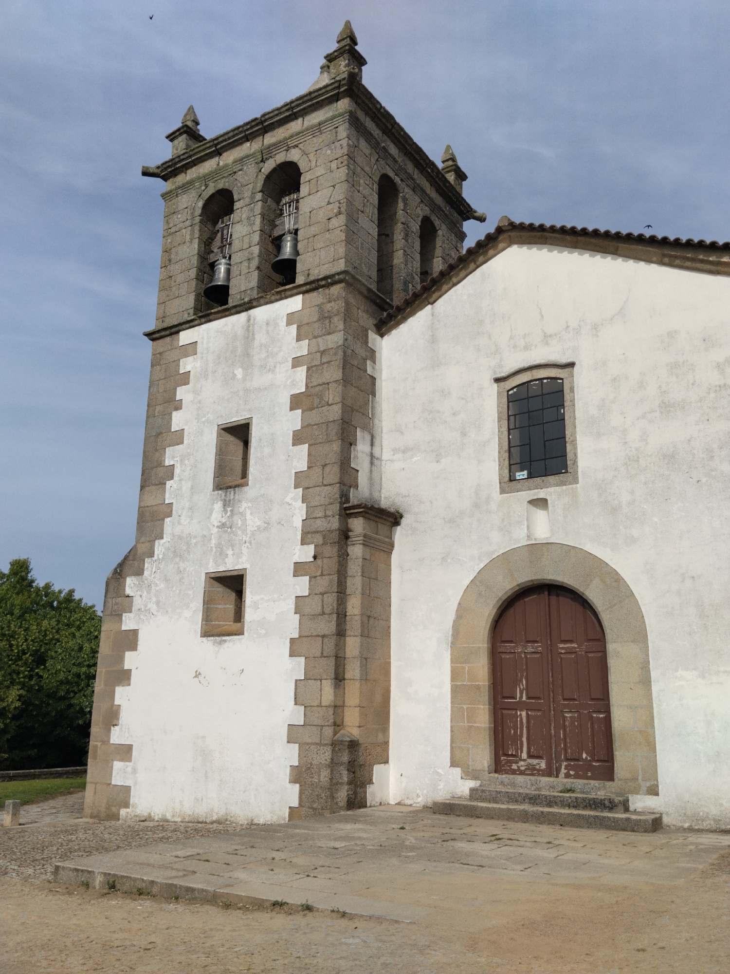 The image depicts a stone church with a bell tower in Mogadouro, Portugal. The church has a white exterior with a stone trim. The bell tower is made of stone and has two visible bells. The church has a single arched wooden door. The surrounding area is paved with stone and dirt. There are trees visible in the background. The sky is overcast.